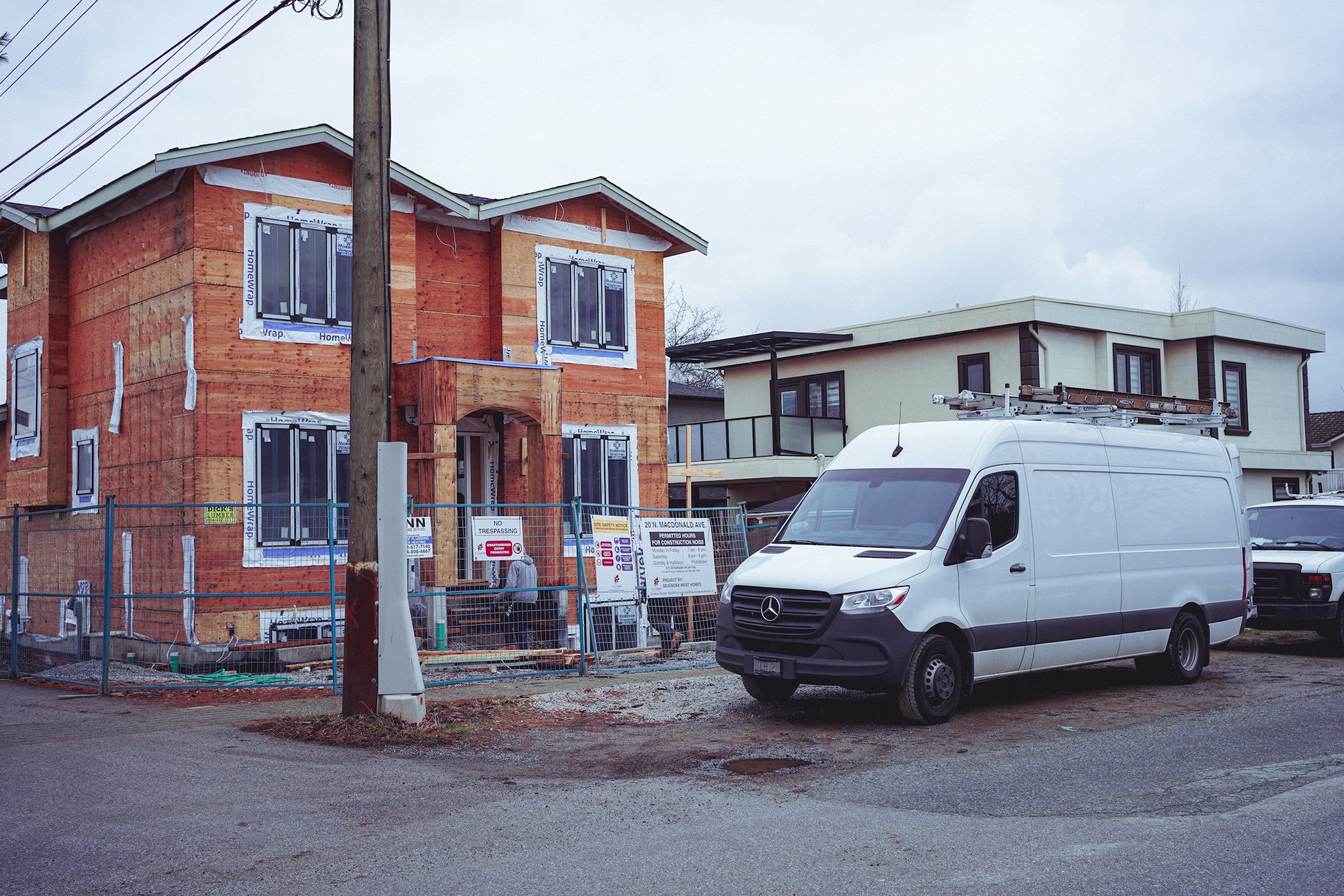 A house under construction with a wooden frame and a white modern neighboring house in a residential area, with a white Mercedes-Benz utility van parked in the foreground and construction signs and fencing around the site.