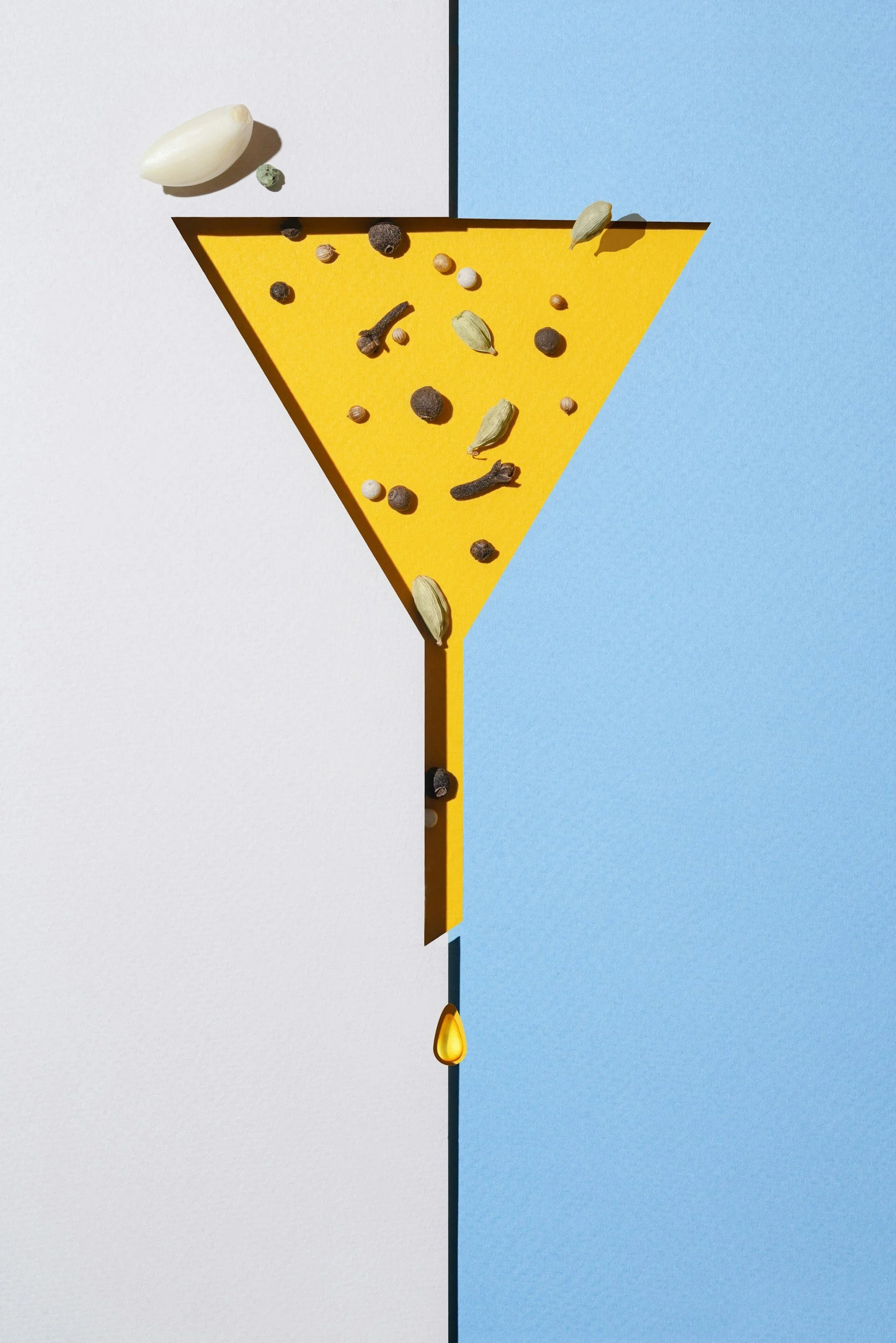 A yellow cocktail glass silhouette with various spices like peppercorns, cardamom pods, and cloves, spilling over the rim and down the stem, with a drop of oil at the bottom, set against a divided blue and white background.