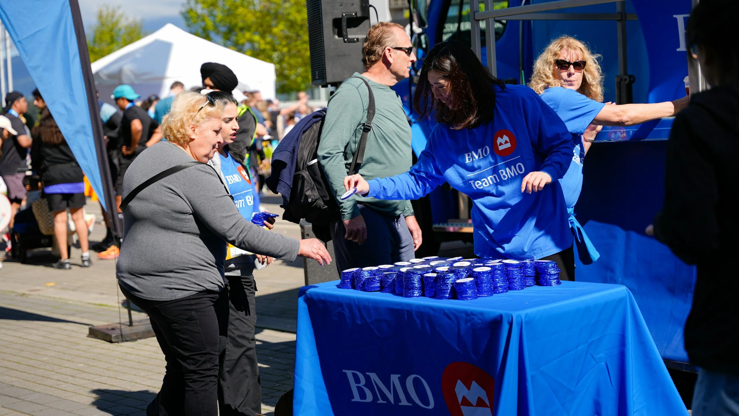 People gather around a table with stacks of medals at an outdoor event, with some handing out medals and others receiving them, under a blue tent with the BMO logo.
