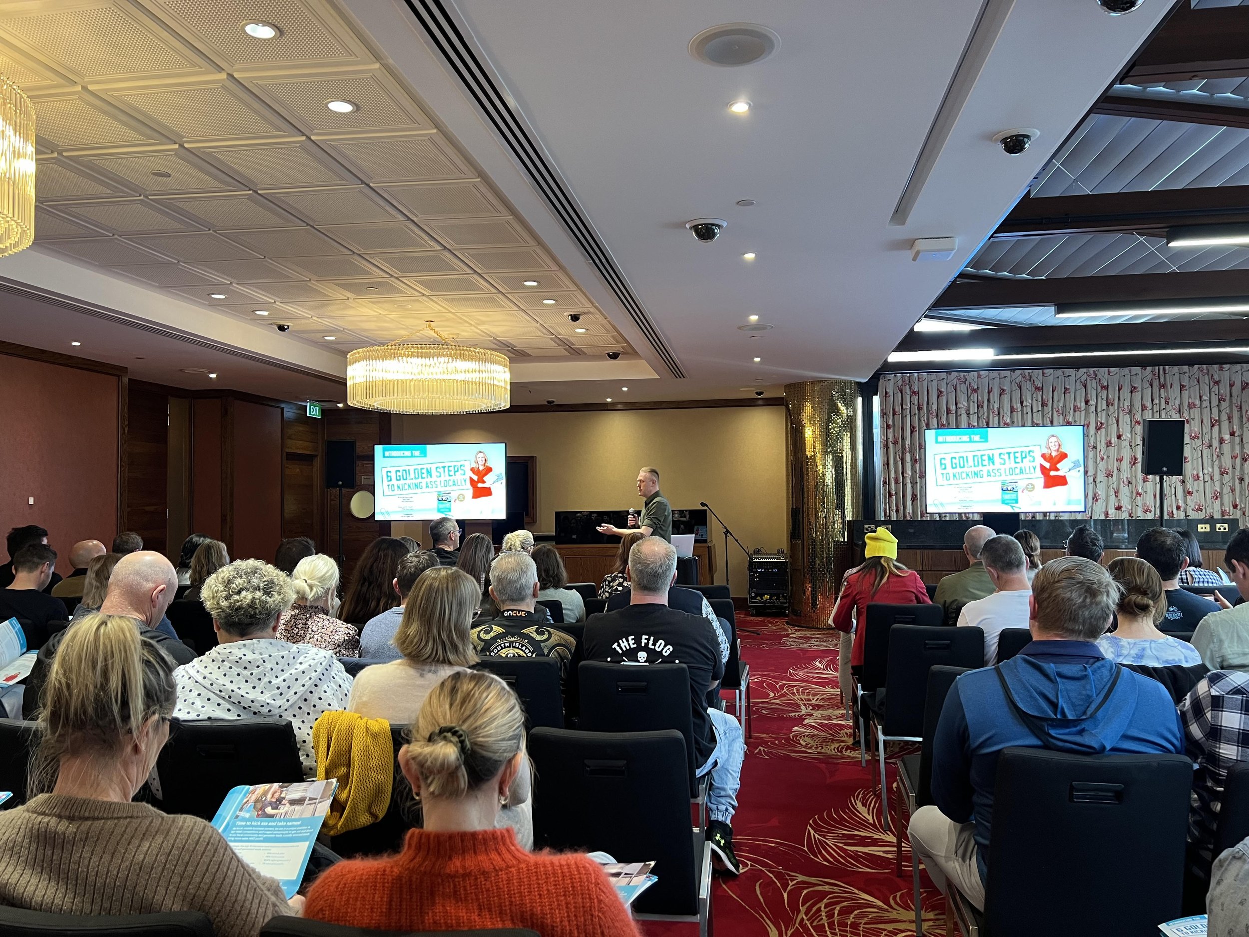 A conference room filled with people sitting in chairs, facing a speaker standing at the front. Two large screens display the presentation title '6 GOLDEN STEPS TO KICKING ASS LOCALLY' with a woman illustration.