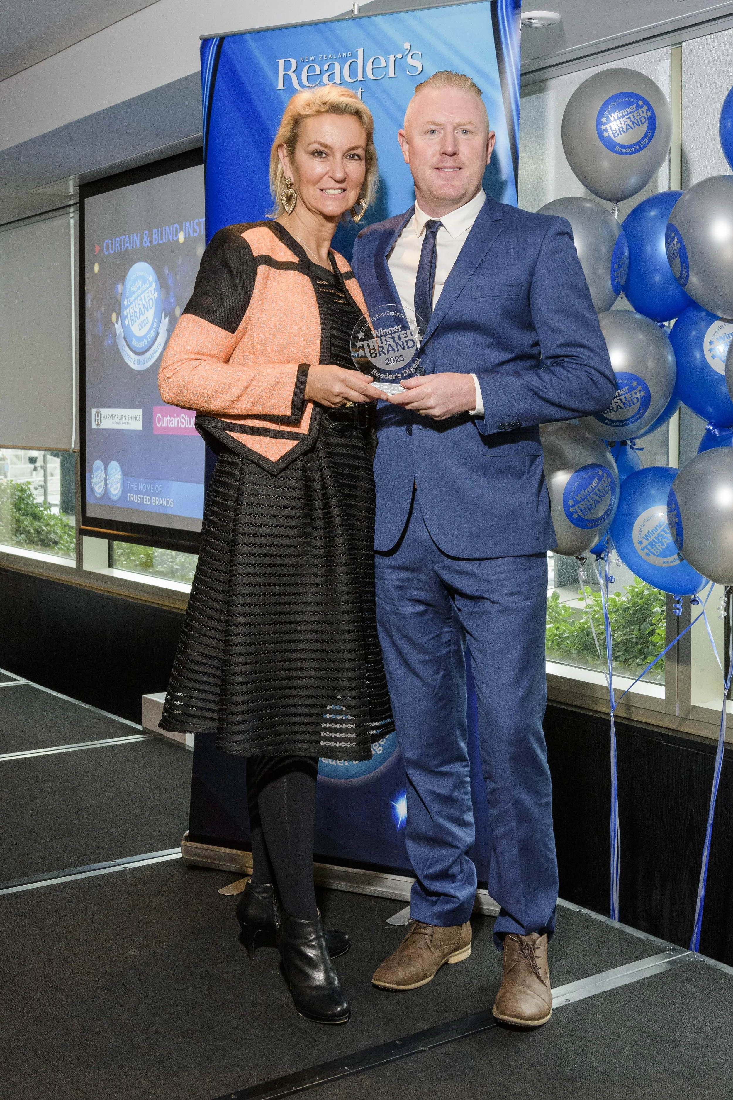 A woman and a man are standing together at an award ceremony, holding a glass trophy. The woman is wearing a black and peach dress with a black textured skirt, and the man is in a blue suit with a white shirt and tie. The background features a large blue banner that reads 'Reader's Digest' and a group of silver and blue balloons with 'Trusted Brand' logos.