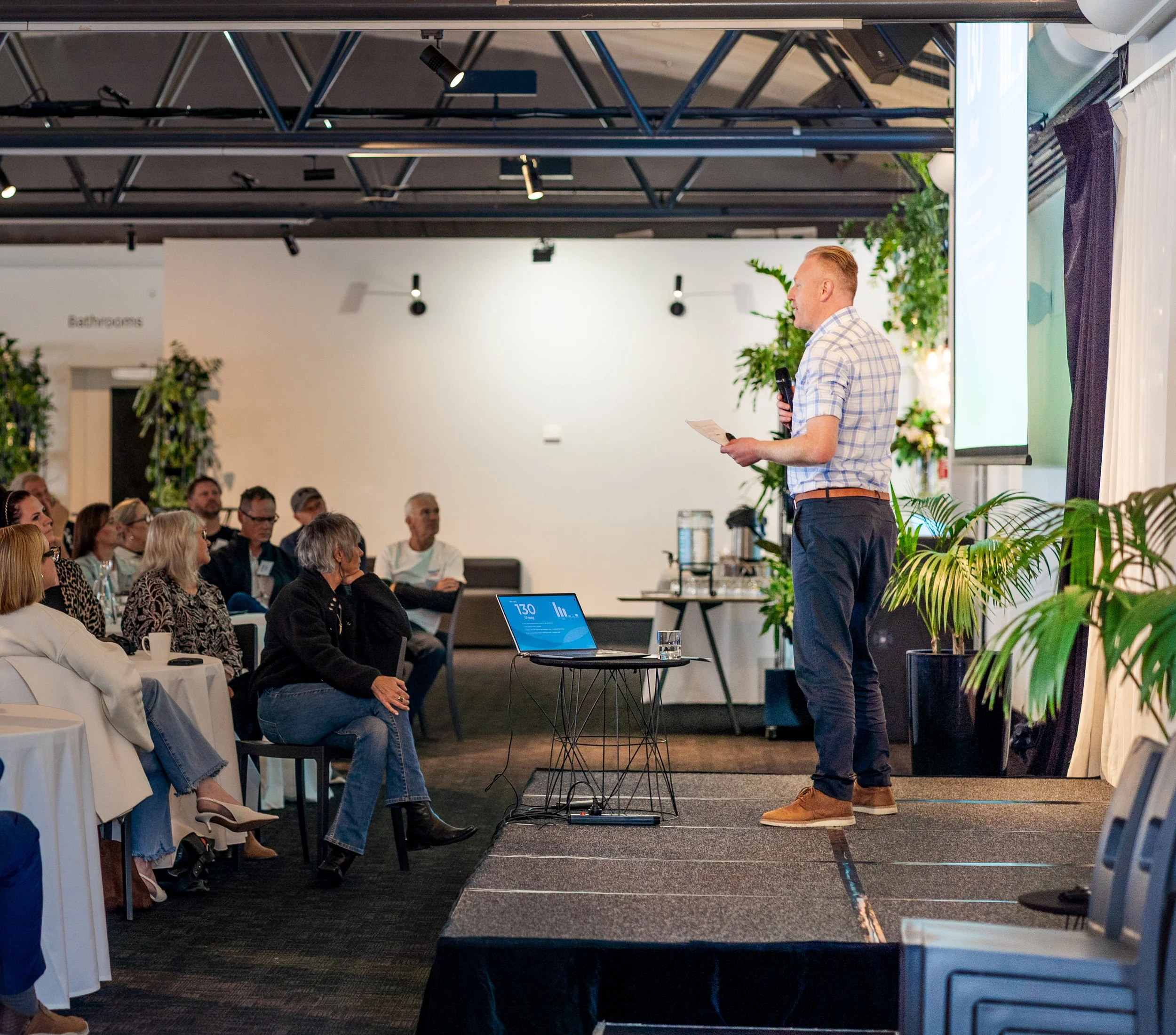 Mitch Grant giving a presentation on a stage with a microphone in a conference room filled with attendees.