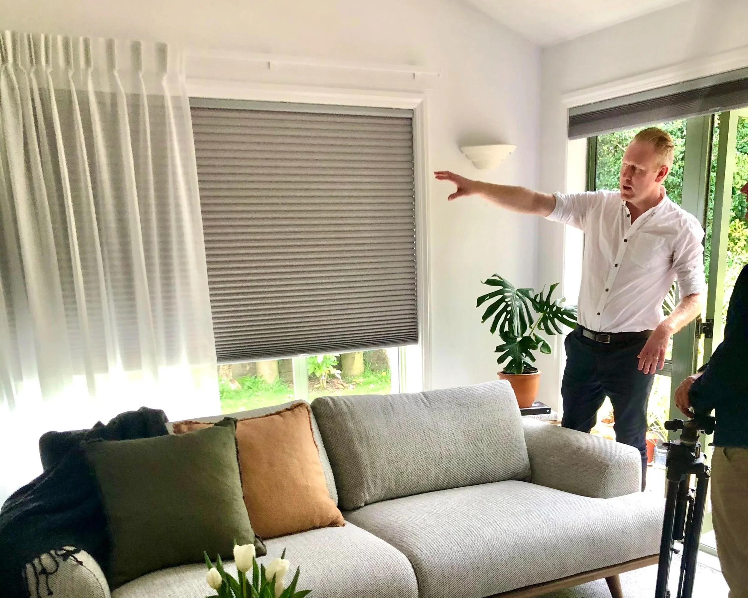A man in a white shirt is gesturing with his right hand while speaking, standing in a living room with a beige sofa, pillows, a potted plant, and a large window with blinds.