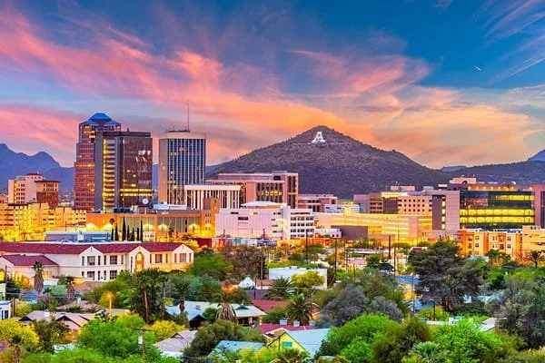 City skyline with modern buildings, green trees, and a mountain with a large letter A on it during sunset.