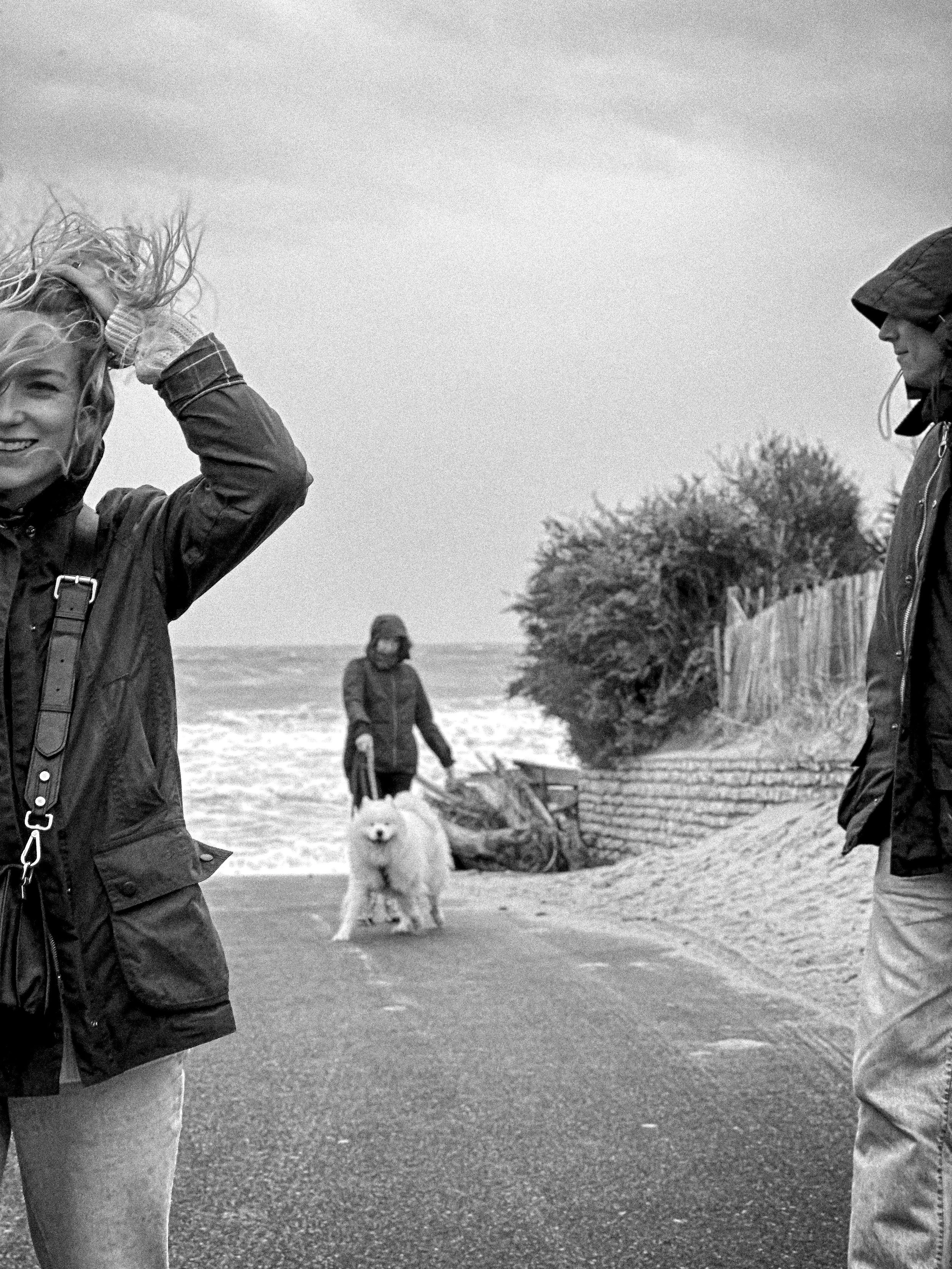 Two people by the sea with wind in their hair and a white dog behind them, Île de Ré, France. Black and white. Photography by Thomas Dohm.