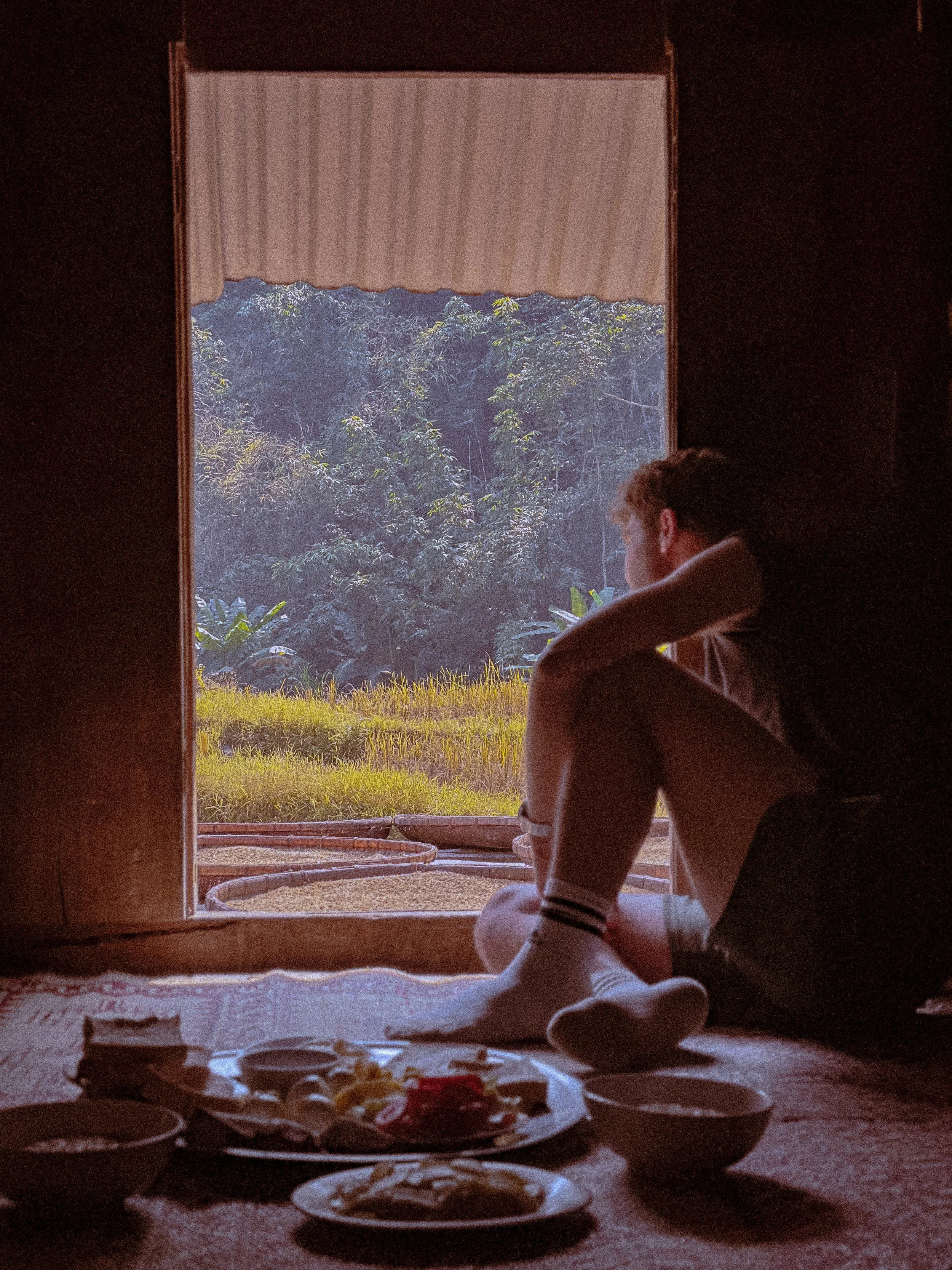 My brother's silhouette seated in a doorway looking out at rice paddies with breakfast laid out, Pu Luong, Vietnam. Photography by Thomas Dohm.