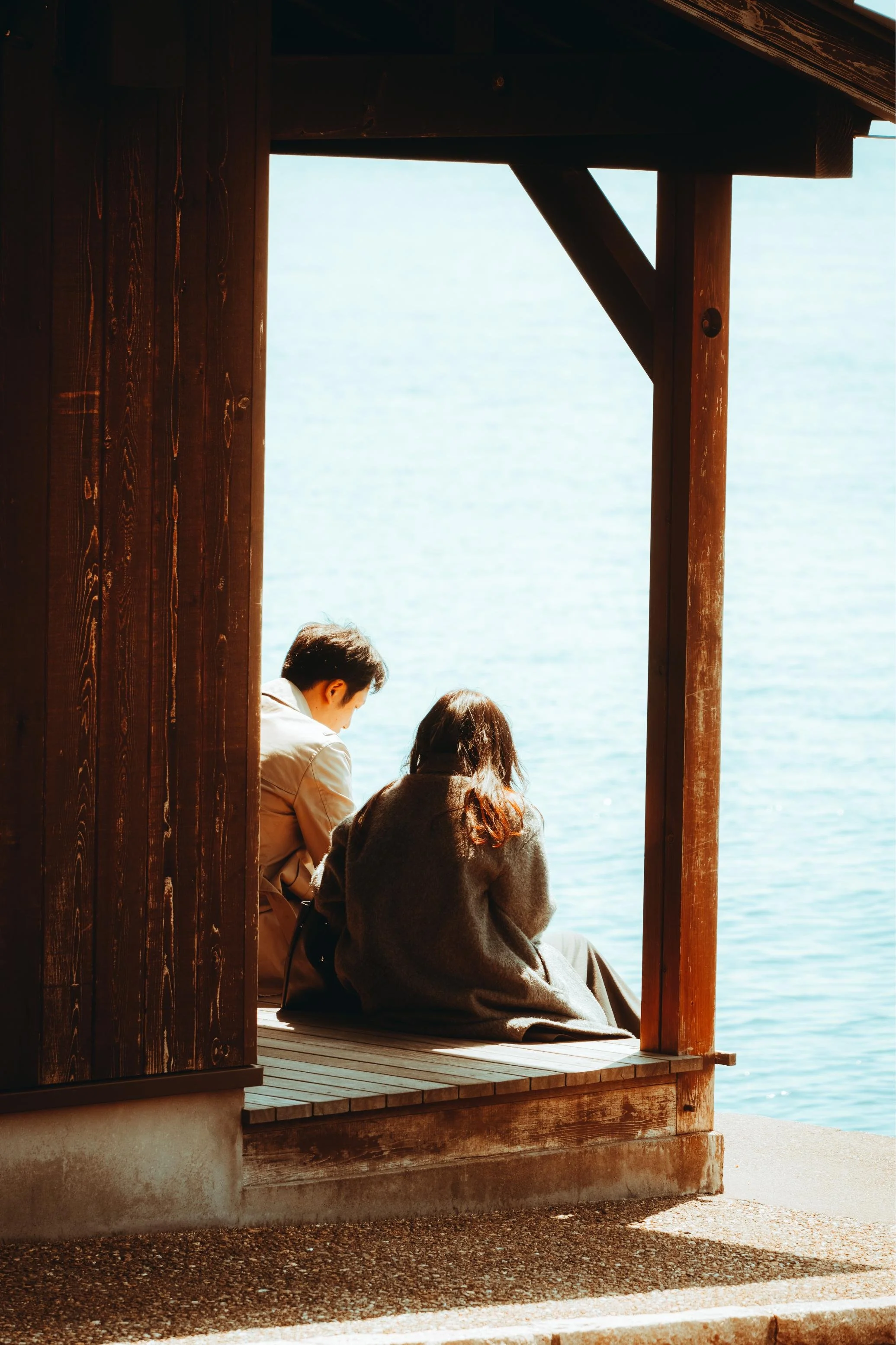 Three people sitting on a wooden deck overlooking a body of water, framed by a wooden structure with a window opening.