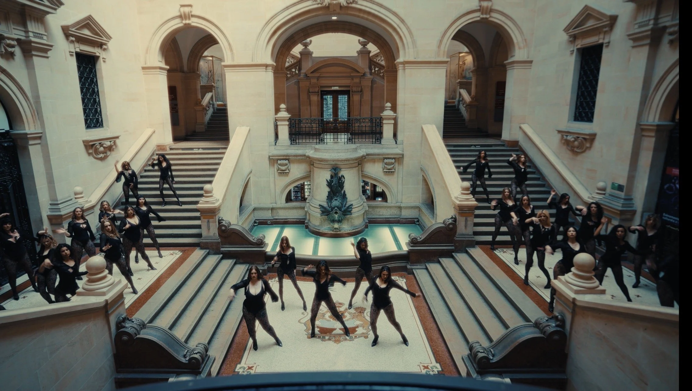 A group of women in black outfits and patterned leggings dancing on the stairs and floor of a grand, historic building with ornate architecture and a water fountain in the center.