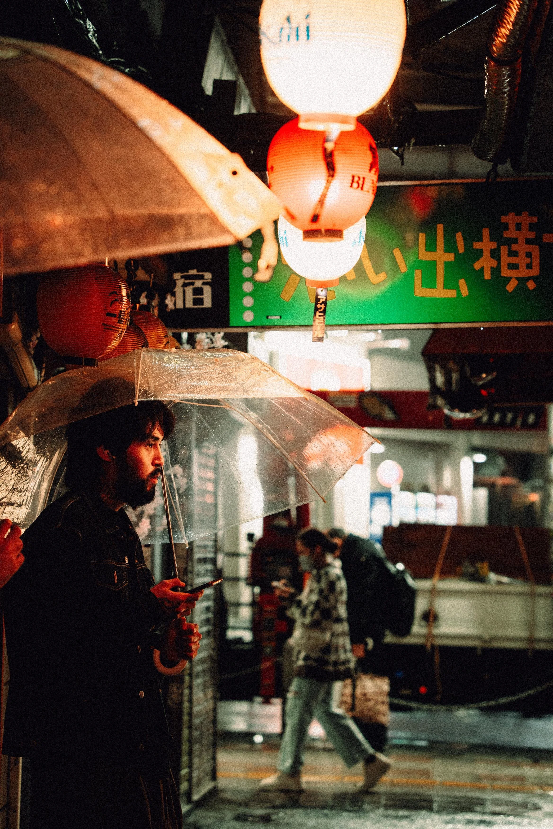 A man with a beard and dark hair holding an umbrella in a rainy urban setting, looking at his phone. There are glowing lanterns hanging, and a green sign with Japanese writing in the background. Other pedestrians are walking past in the rain.