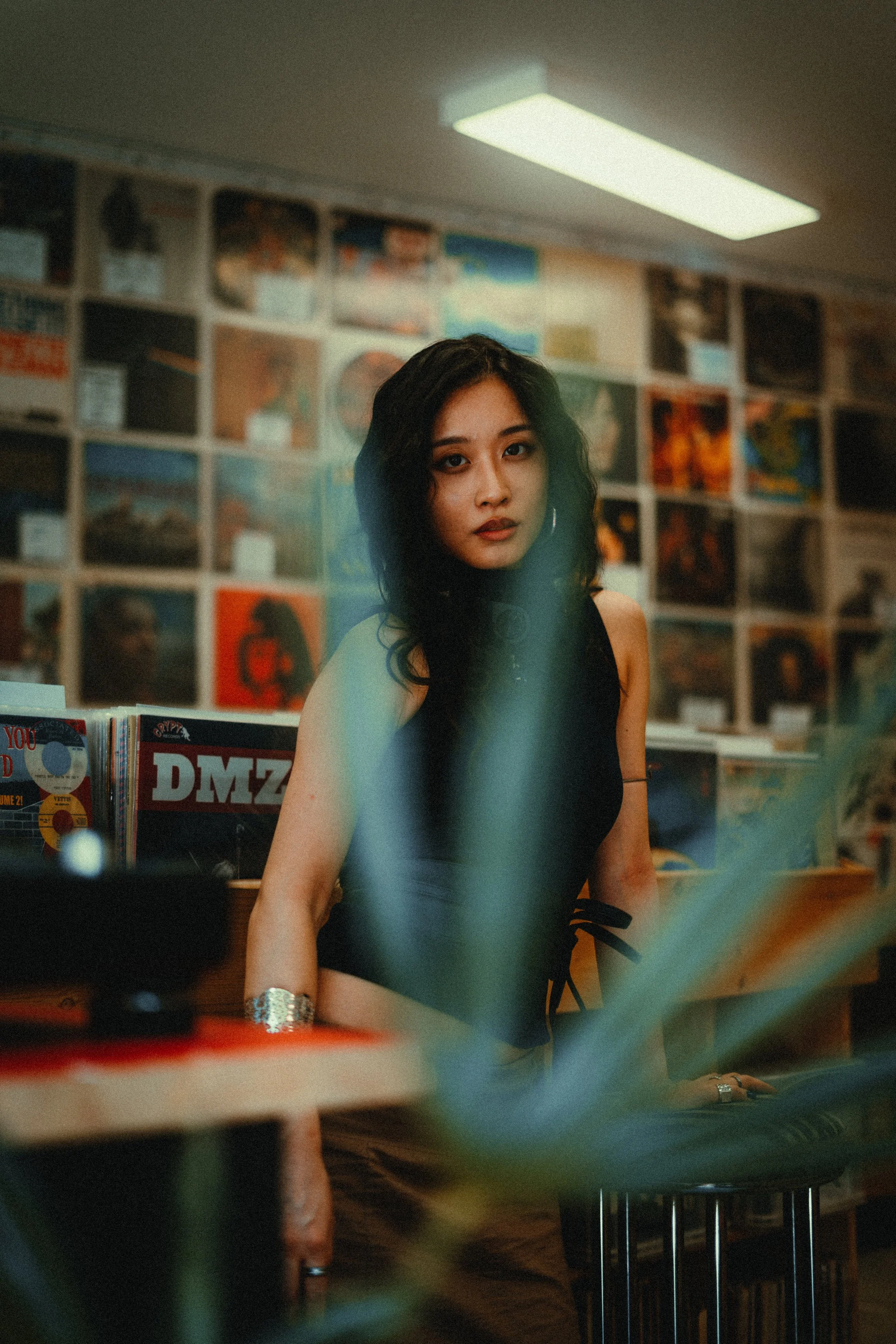 A young woman with dark, wavy hair and hoop earrings, dressed in a black top, is sitting at a wooden counter in a record store. The background features a colorful wall of album covers, and the scene is softly lit with overhead lighting.