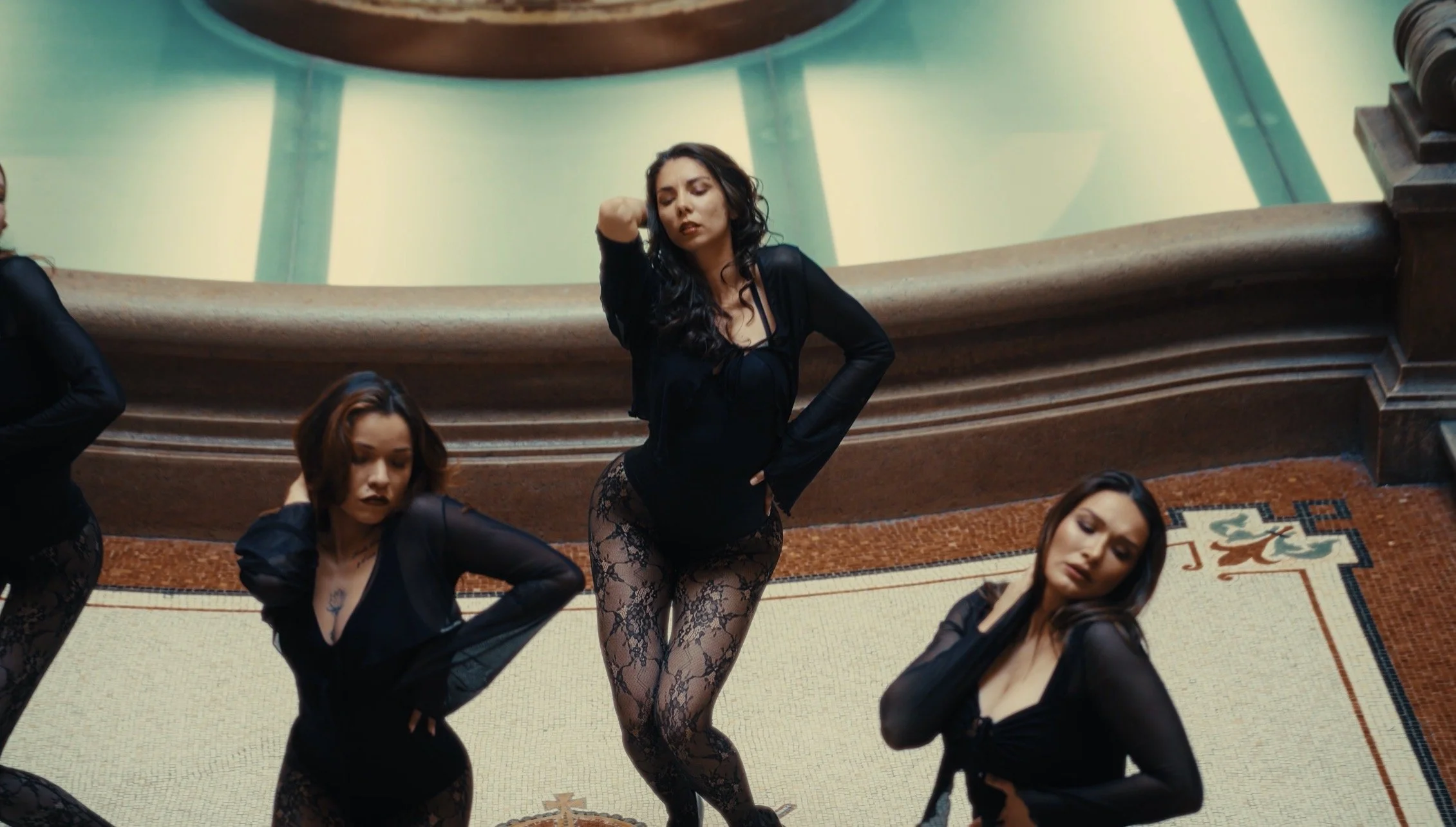 Group of women dressed in black dance costumes with lace tights, performing in front of a mosaic in an ornate theater or opera house.