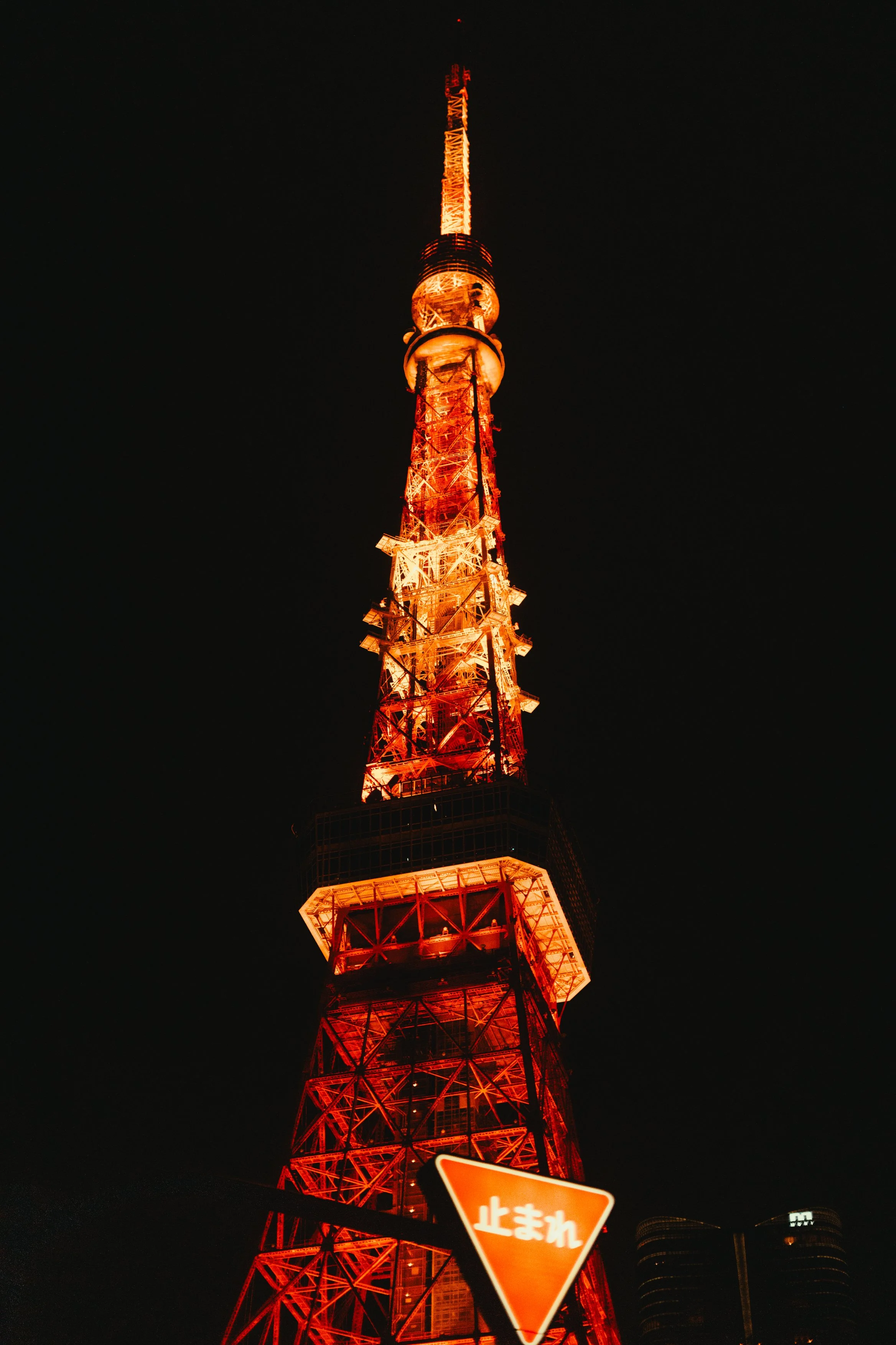 Night view of Tokyo Tower illuminated in orange. The tower has a lattice structure and a large observation deck. A Japanese stop sign with the text '止まれ' is in the foreground.