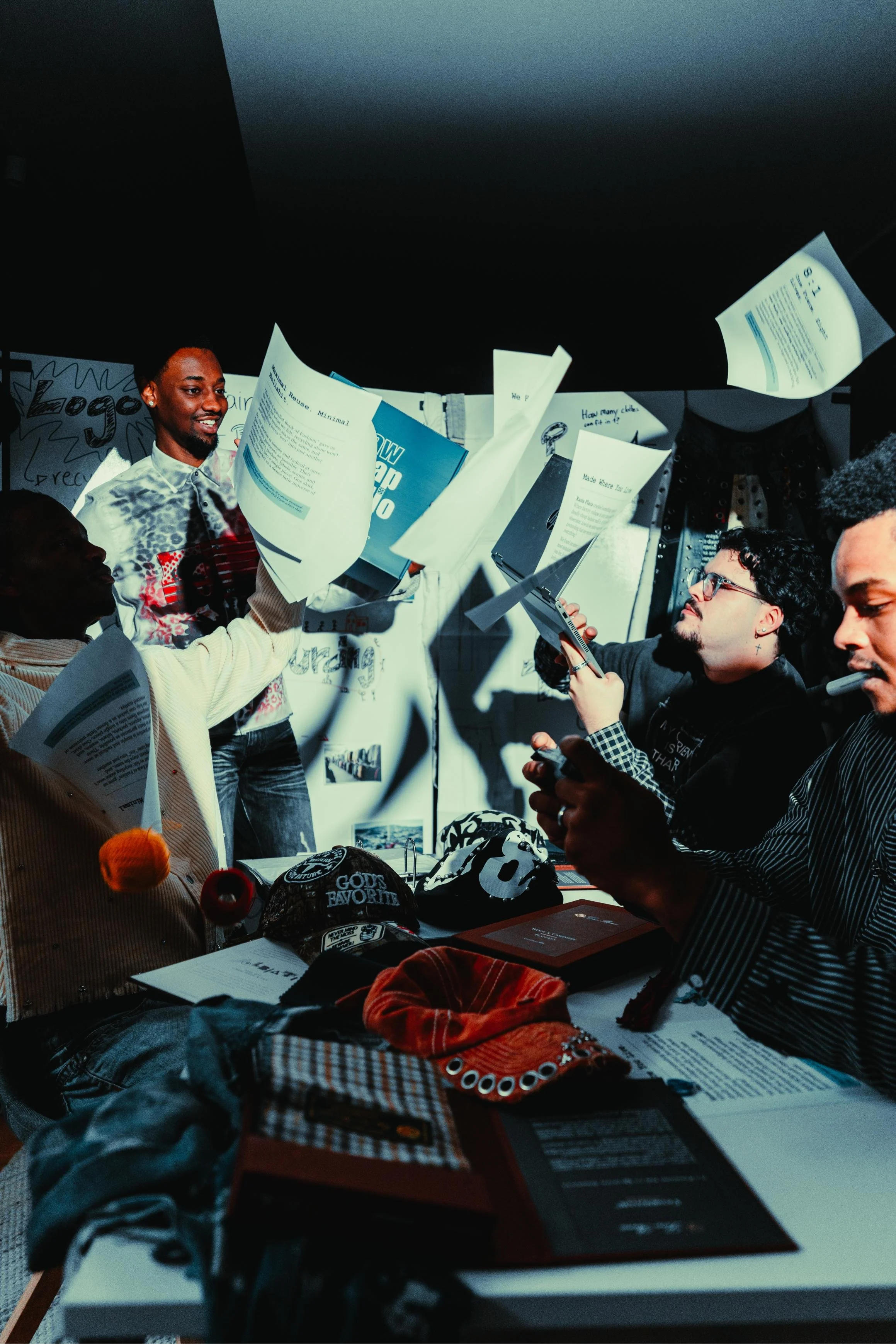 Several people are gathered around a table at an indoor event, looking at papers and possibly discussing or presenting. There are posters and signs displayed on the wall behind them, with one partially visible reading 'Logo'. The table has various ob