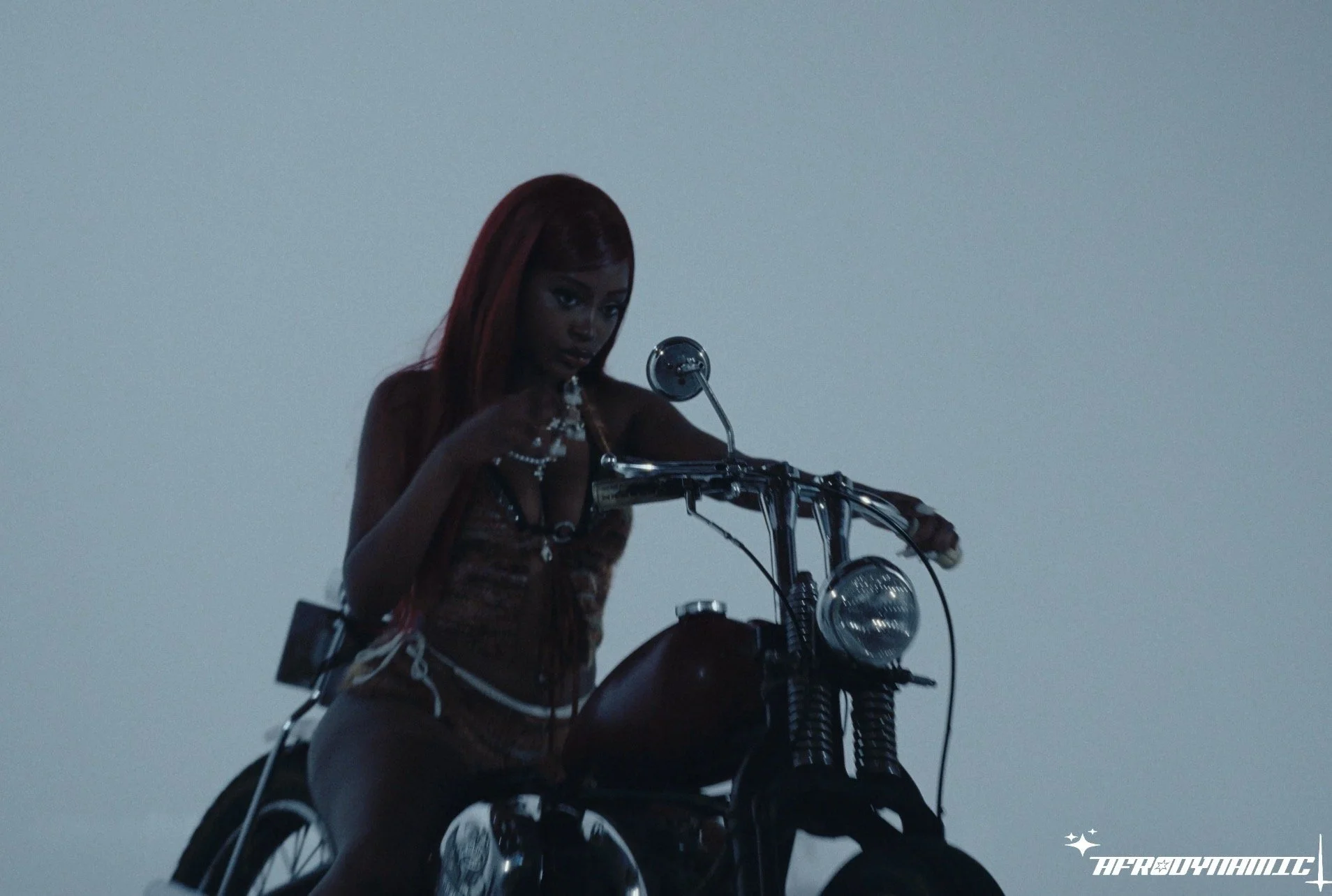 A woman with long red hair wearing jewelry, sitting on a motorcycle against a cloudy sky background.