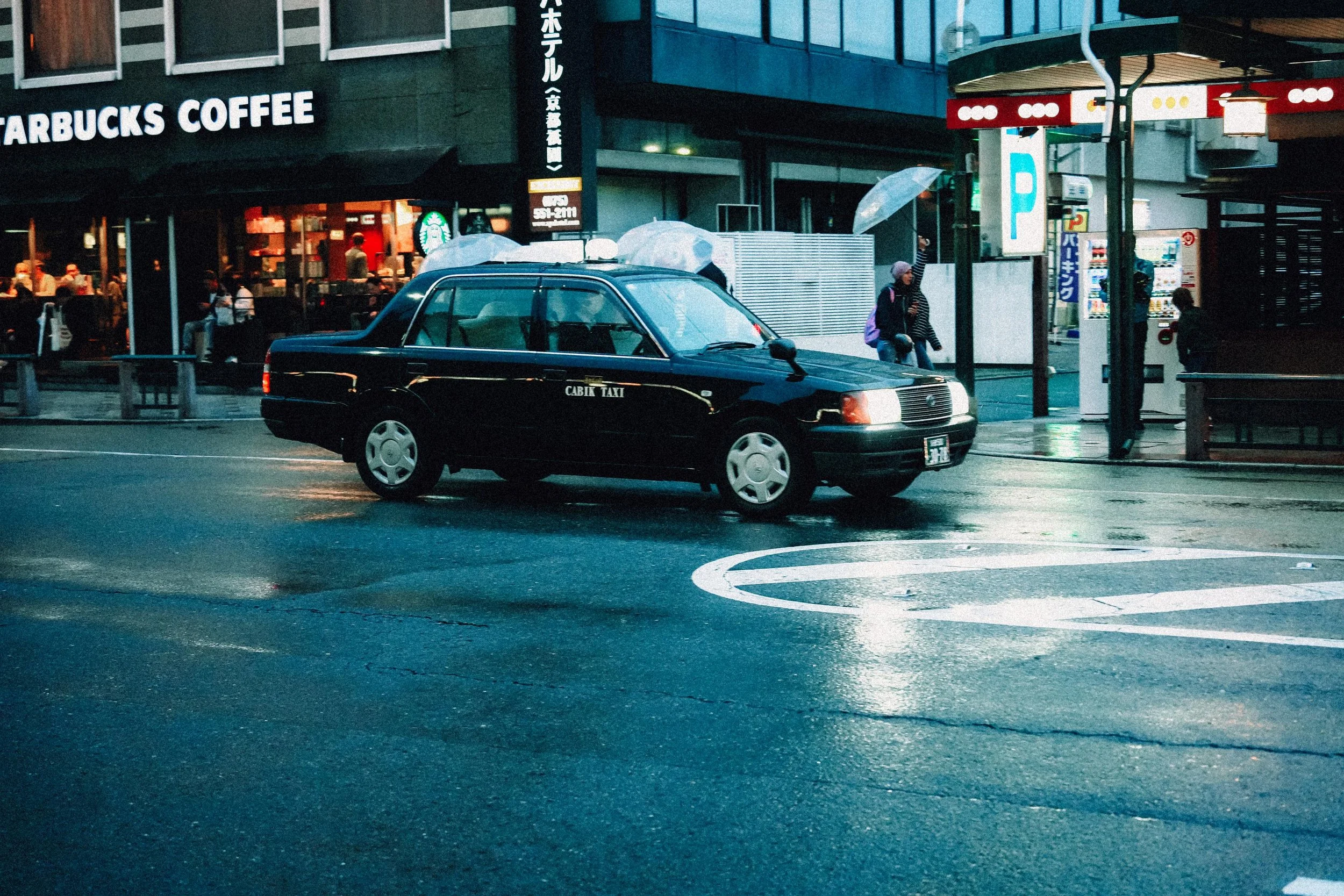 Nighttime street scene featuring a black taxi cab parked near a sidewalk, with a Starbucks Coffee shop and various pedestrians in the background.