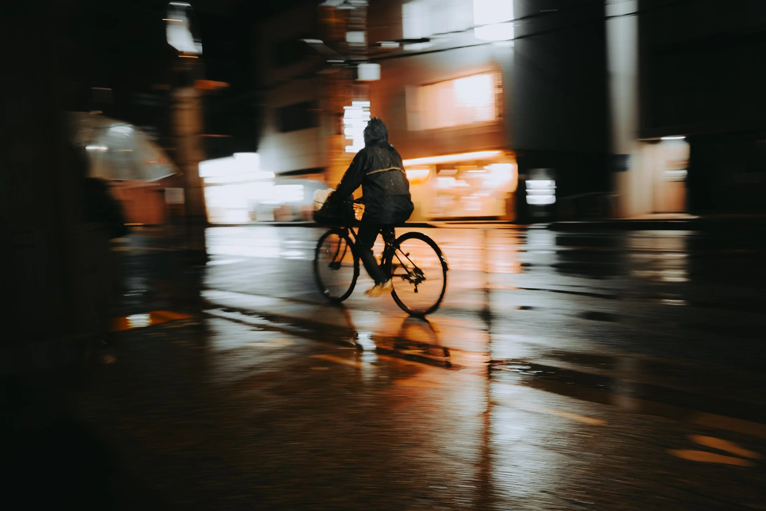 A person riding a bicycle through a wet street at night, with reflections of city lights on the surface, and a blurry background of buildings and lights.