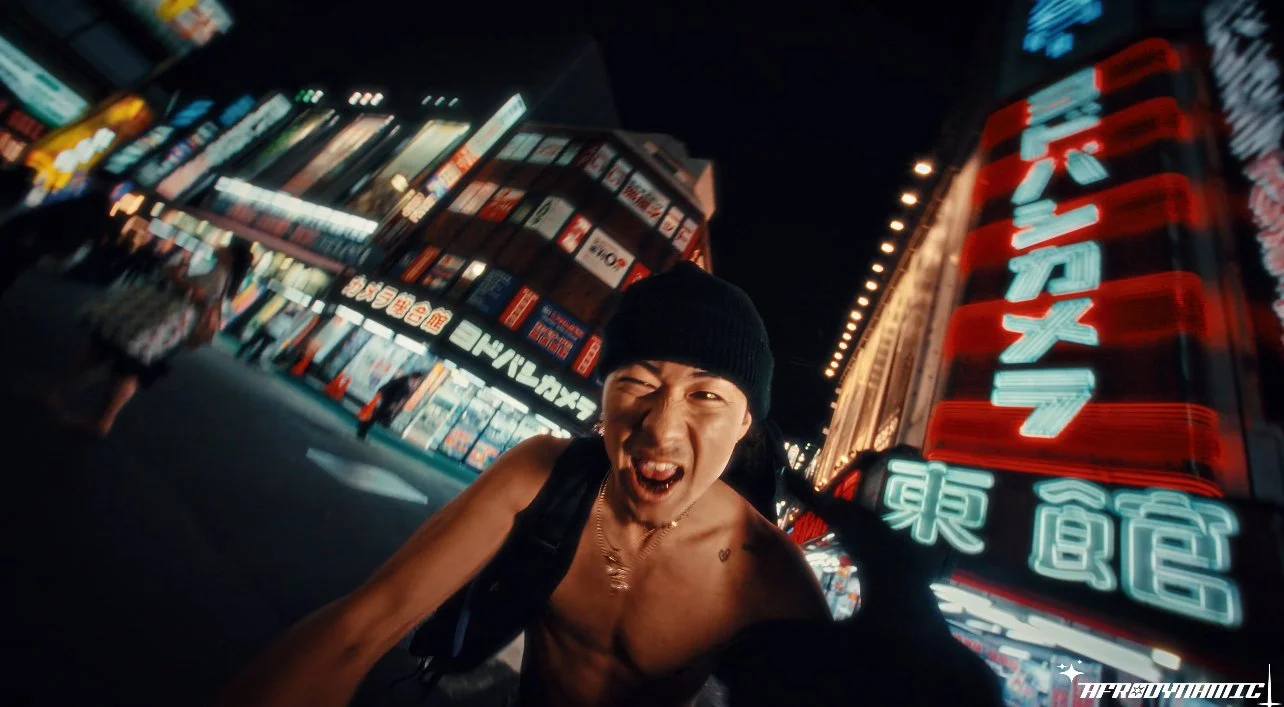 A shirtless young man wearing a black beanie, making a playful face in a city street at night with bright neon signs in the background.