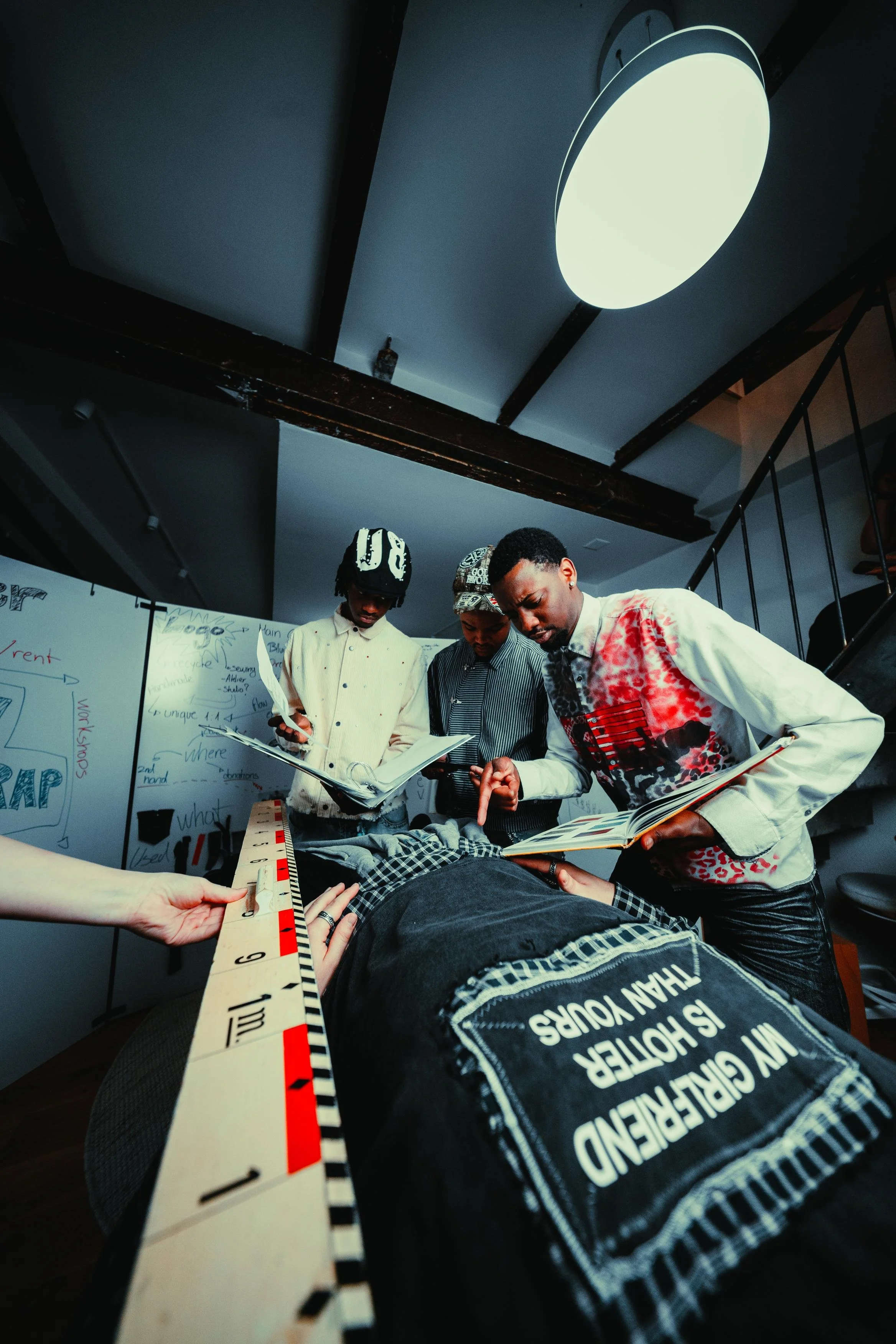 Three young men are gathered around a table, examining books and papers. One man is lying down with his head and upper body on the table, wearing a shirt with a political message. The other two men are standing, one wearing a hat and the other wearin