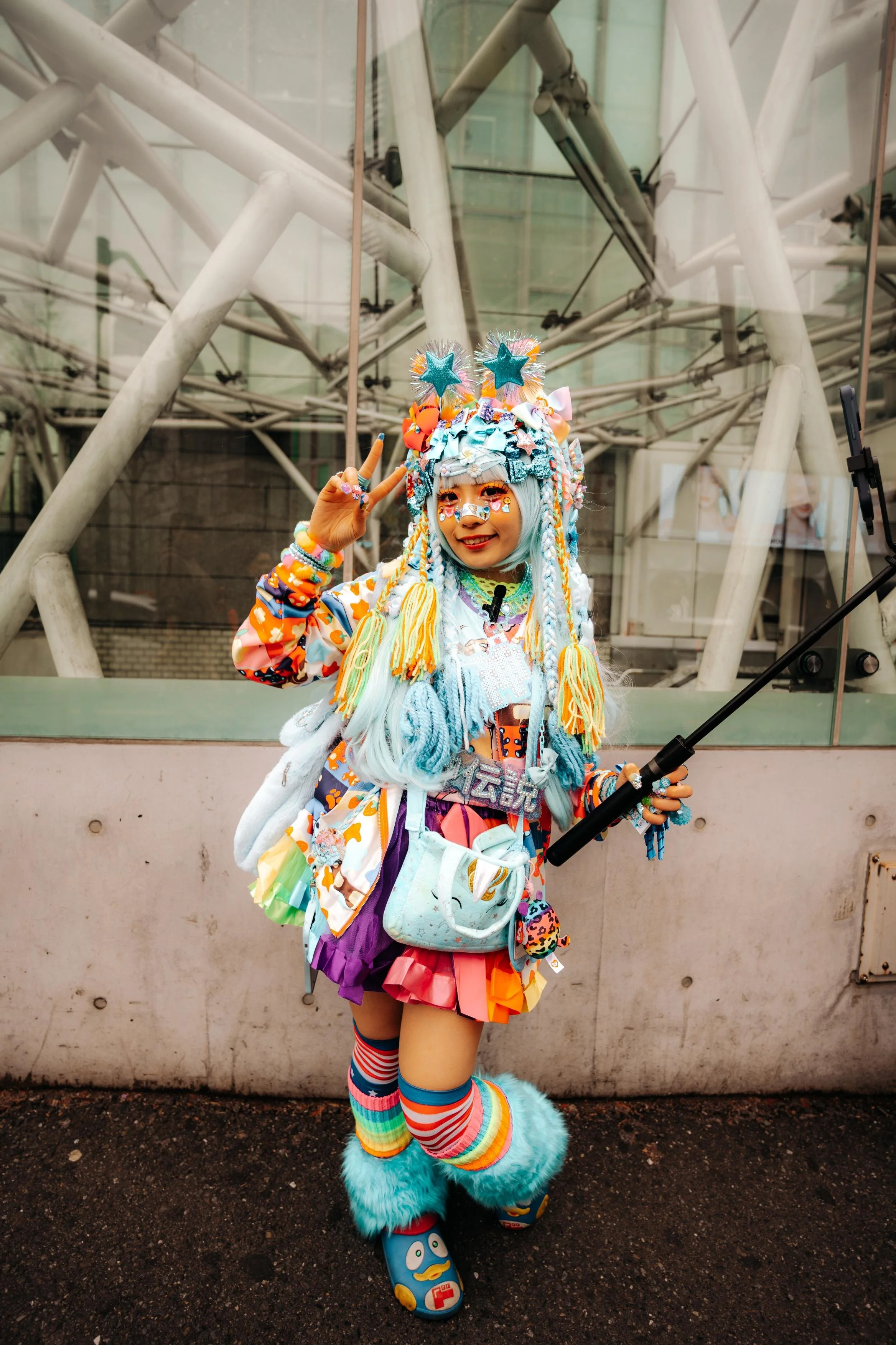 A person dressed in colorful, eccentric fashion, with bright pastel blue hair, making a peace sign and smiling. They are wearing a vibrant, mixed-patterned outfit with accessories, including fluffy teal boots, rainbow knee-high socks, and various pla