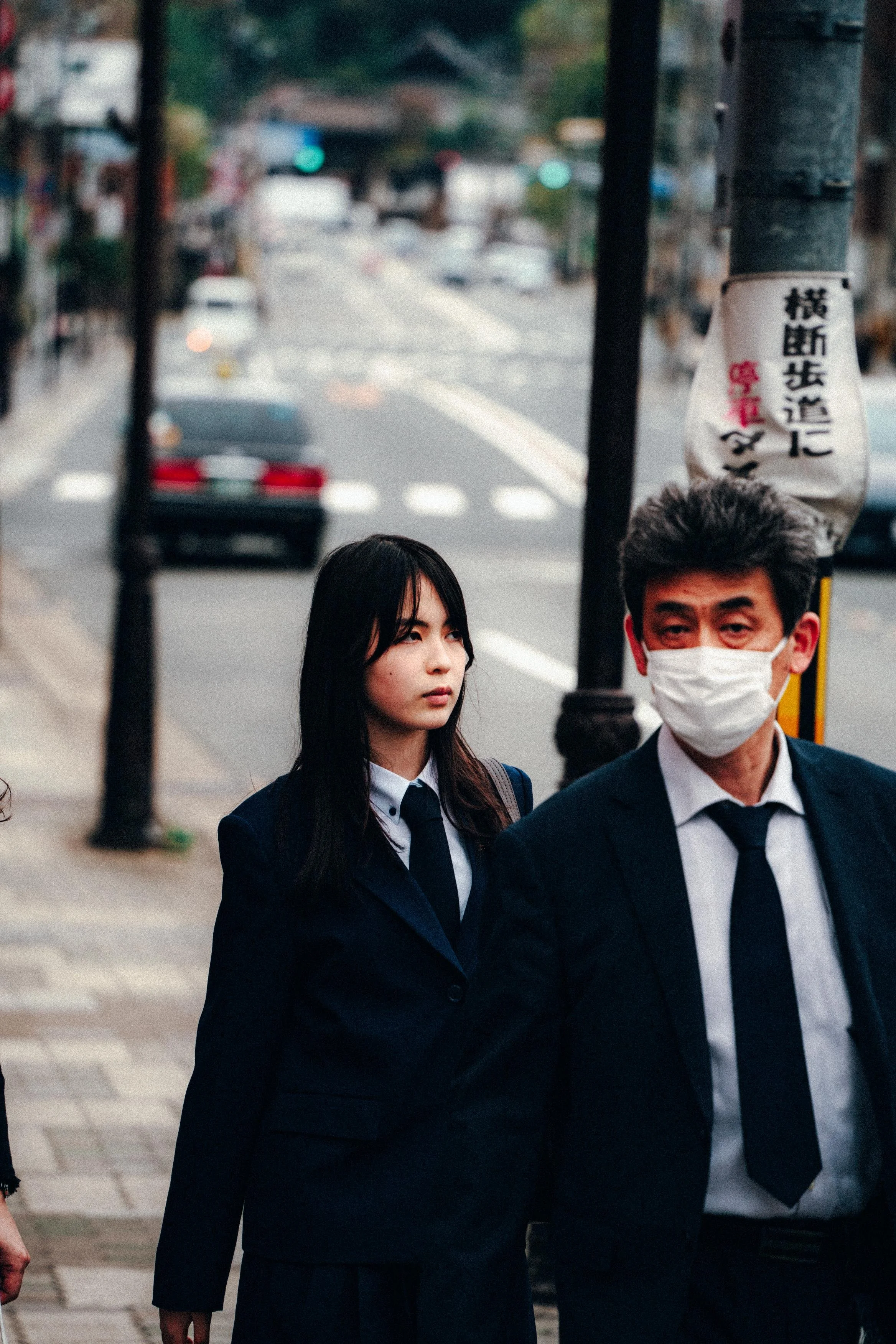 A young woman in a dark suit and tie walks on a city sidewalk alongside an older man wearing a face mask. The background shows a street scene with cars and traffic lights, and some signage in Japanese.