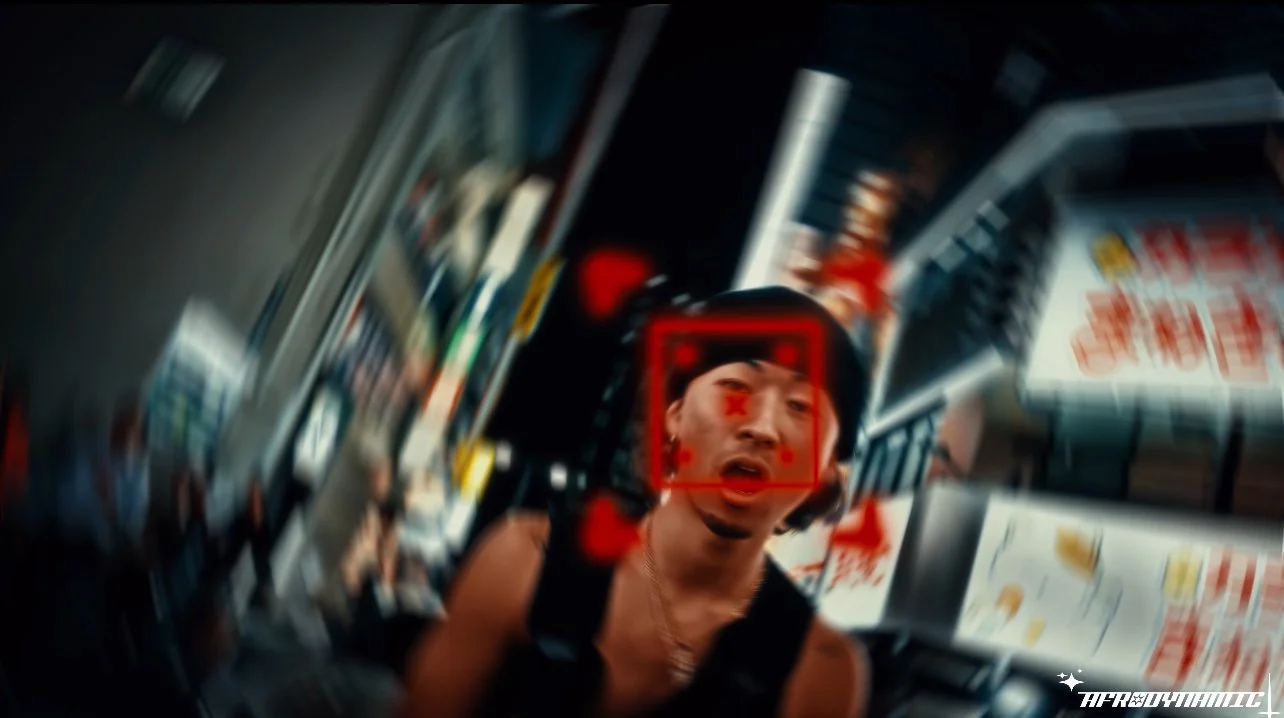 A person with a surprised expression is being captured by a camera's facial recognition system in an urban setting at night, with bright storefront signs and reflections in the background.