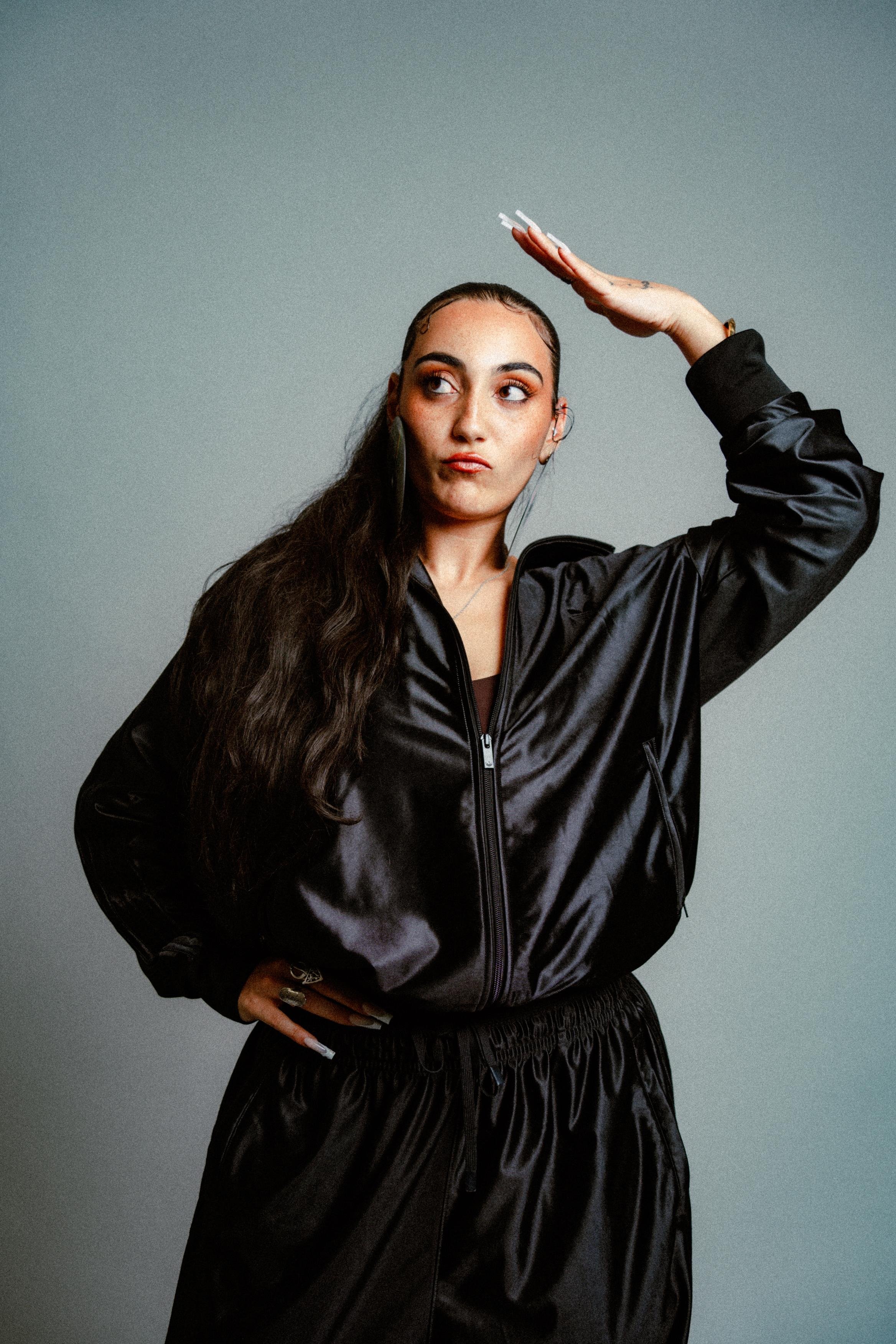 A young woman with long dark hair, dressed in a black shiny jacket and black pants, posing with her right hand raised above her head and her left hand on her hip, against a plain gray background.