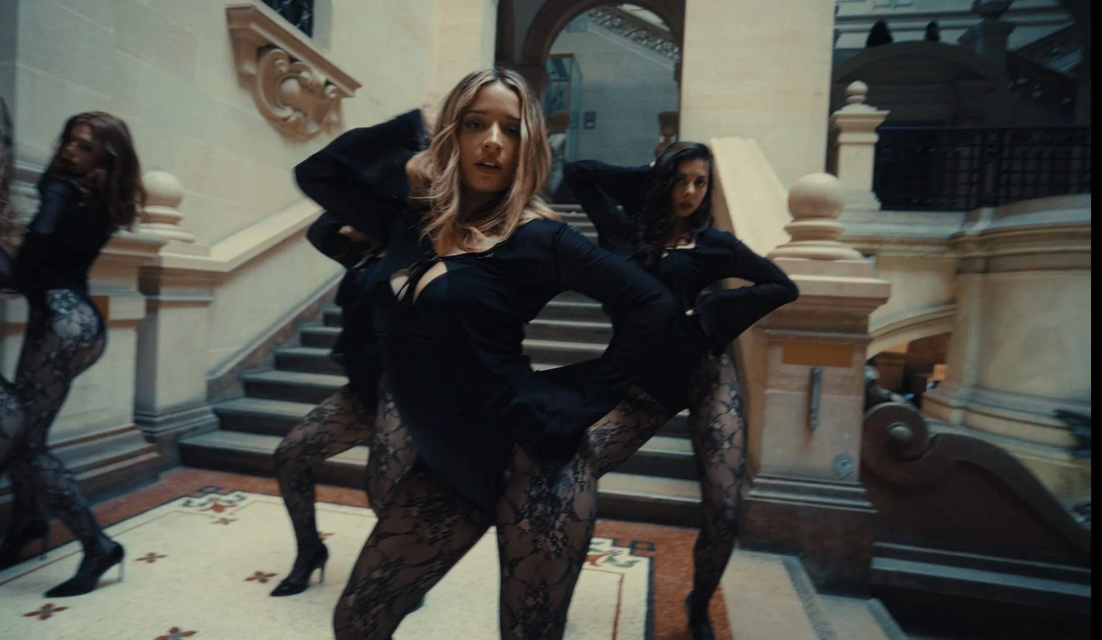 A woman dancing at the top of a staircase inside a grand building with ornate architectural details, accompanied by two women in matching black lace outfits.