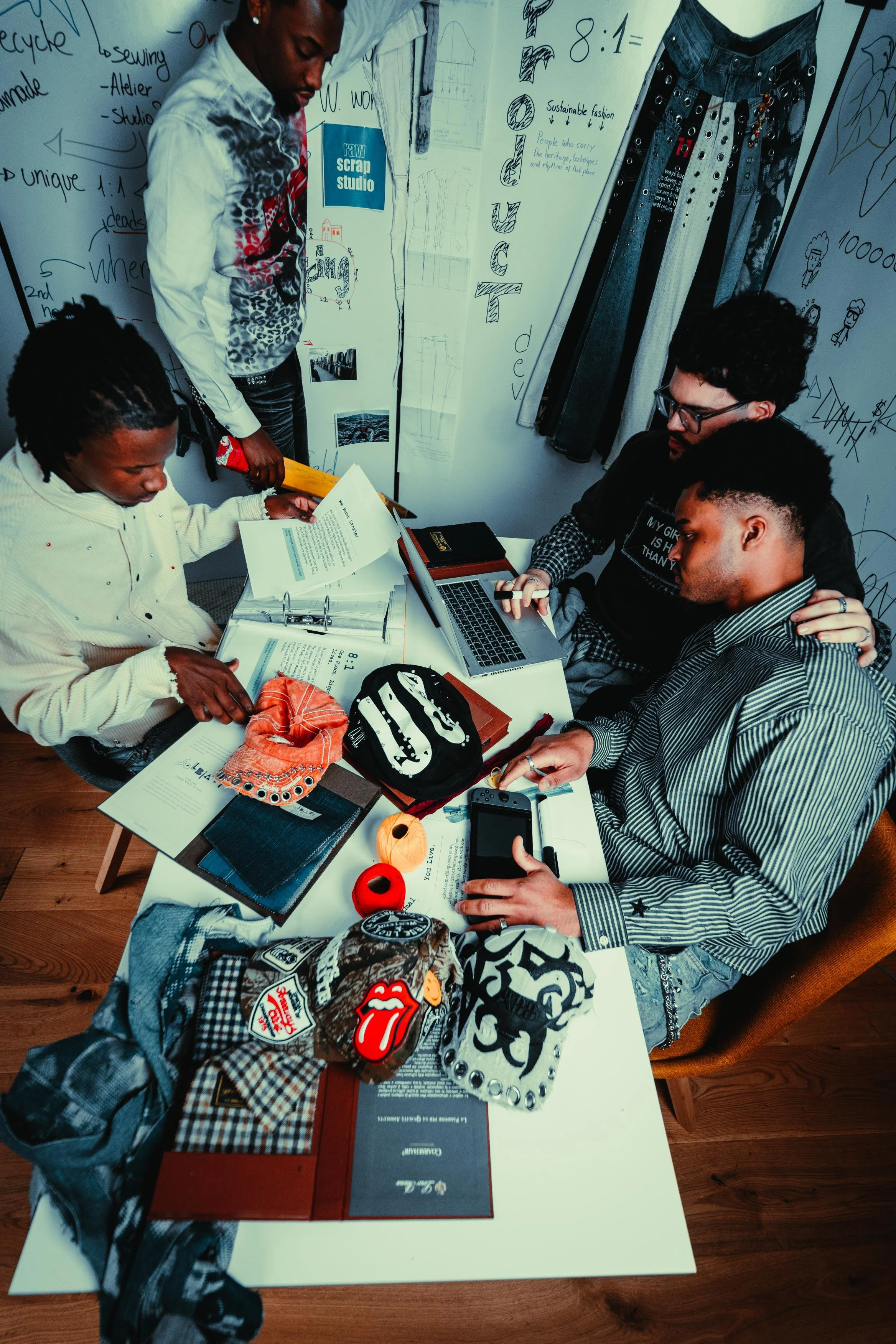 A group of four people gathered around a table in a creative or fashion design workspace, looking at documents, books, and fabric swatches, with whiteboards covered in sketches, notes, and fabric samples on the wall behind them.