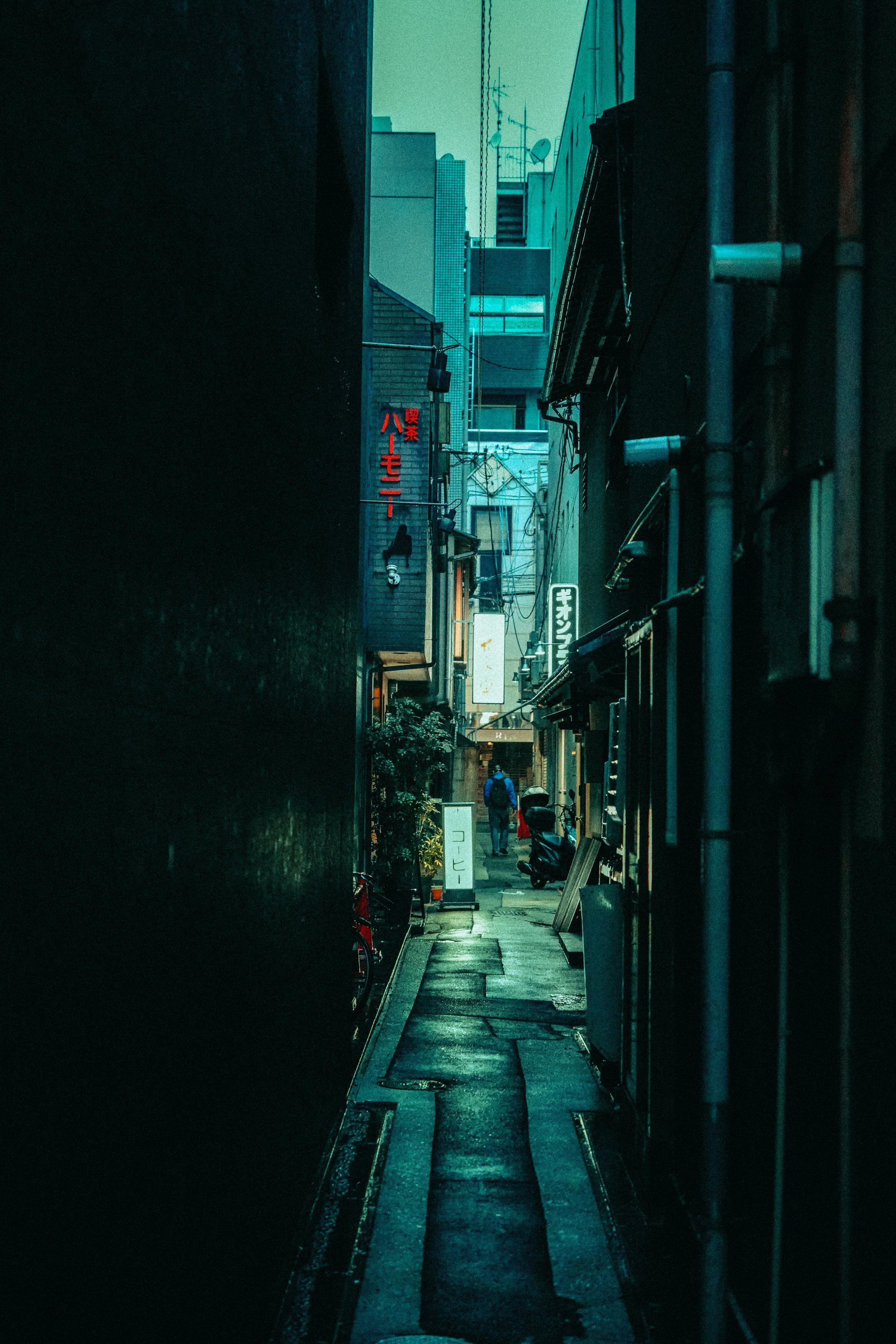 Narrow alleyway of urban street at dusk with a person walking and a scooter parked, illuminated by soft city lights.