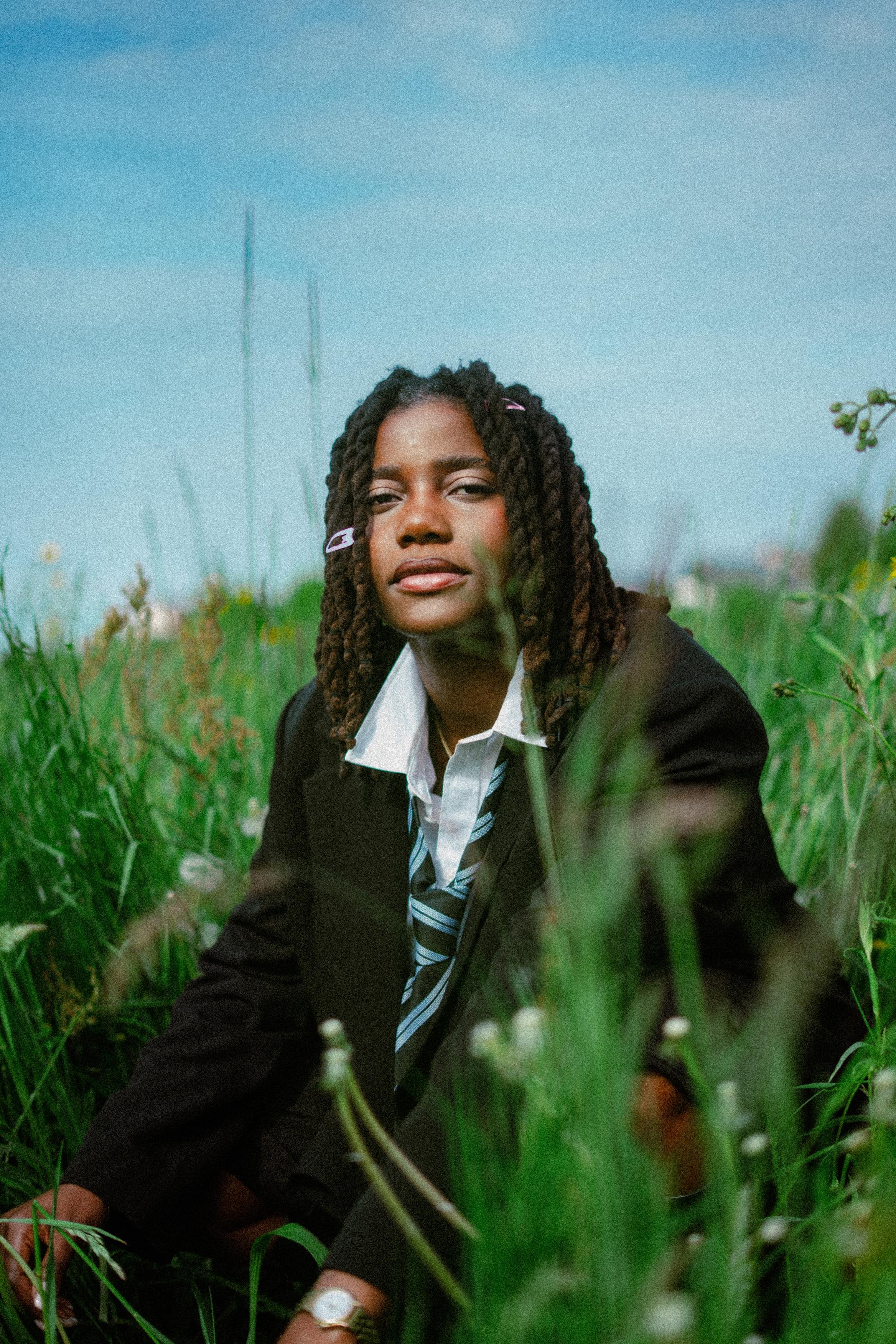 A woman with dreadlocks and a white shirt, wearing a black blazer and a striped tie, sitting in a grassy field with a clear blue sky in the background.