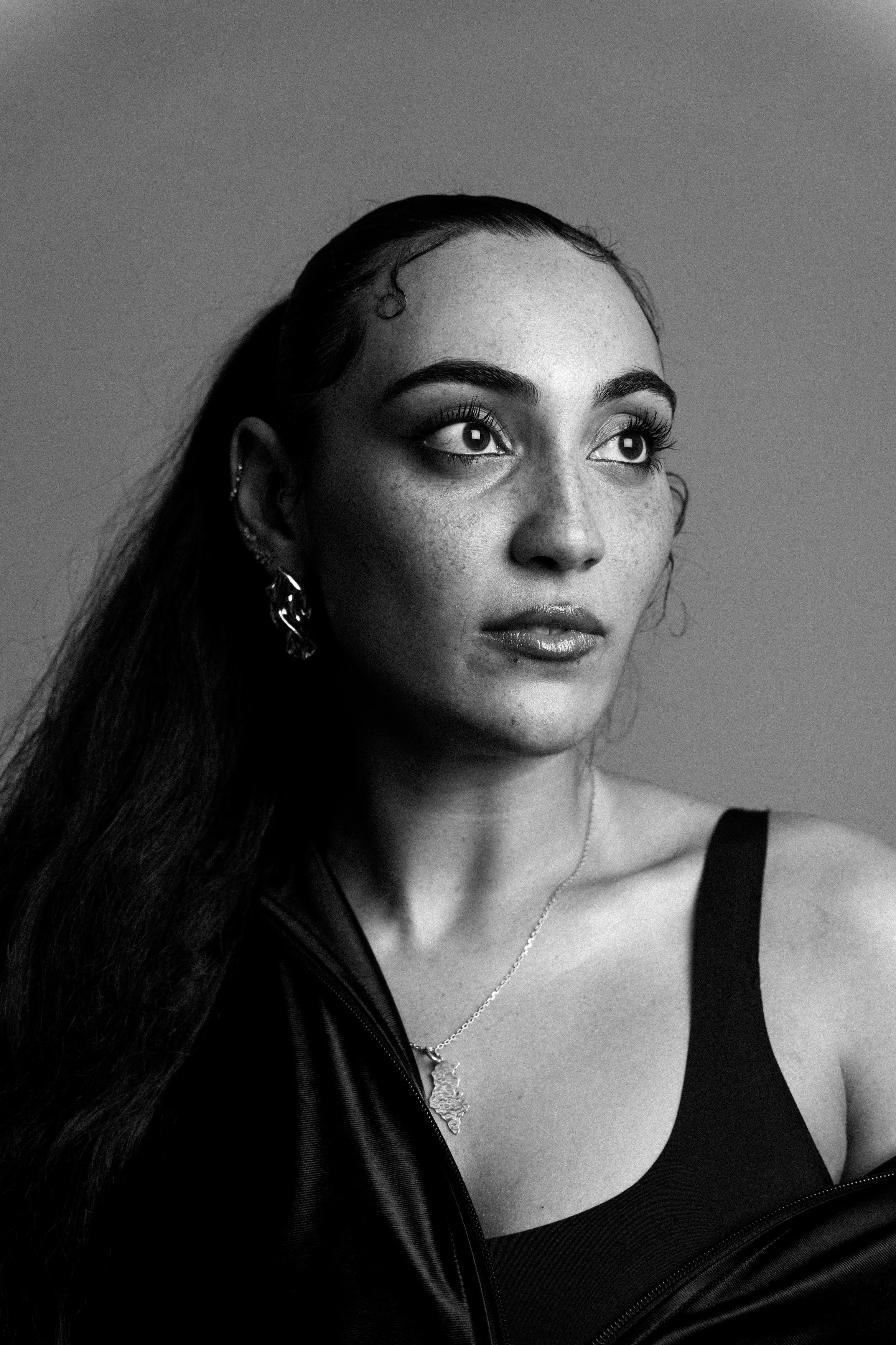 Black and white portrait of a woman with long hair, wearing a tank top, a zip-up jacket, earrings, and a necklace, looking slightly upward.