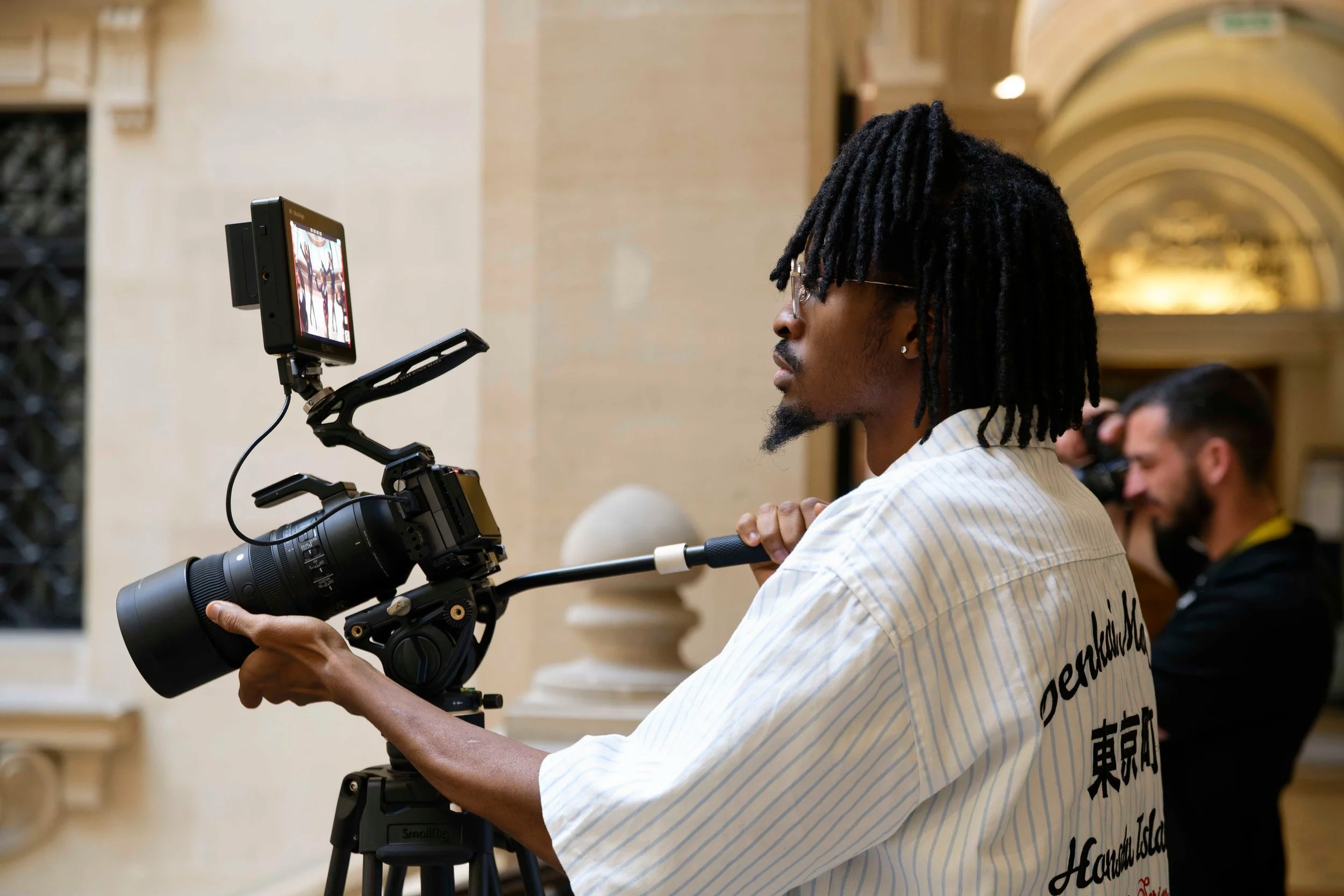 A person with dreadlocks and glasses operating a professional video camera on a tripod in an indoor setting, with another person visible in the background.