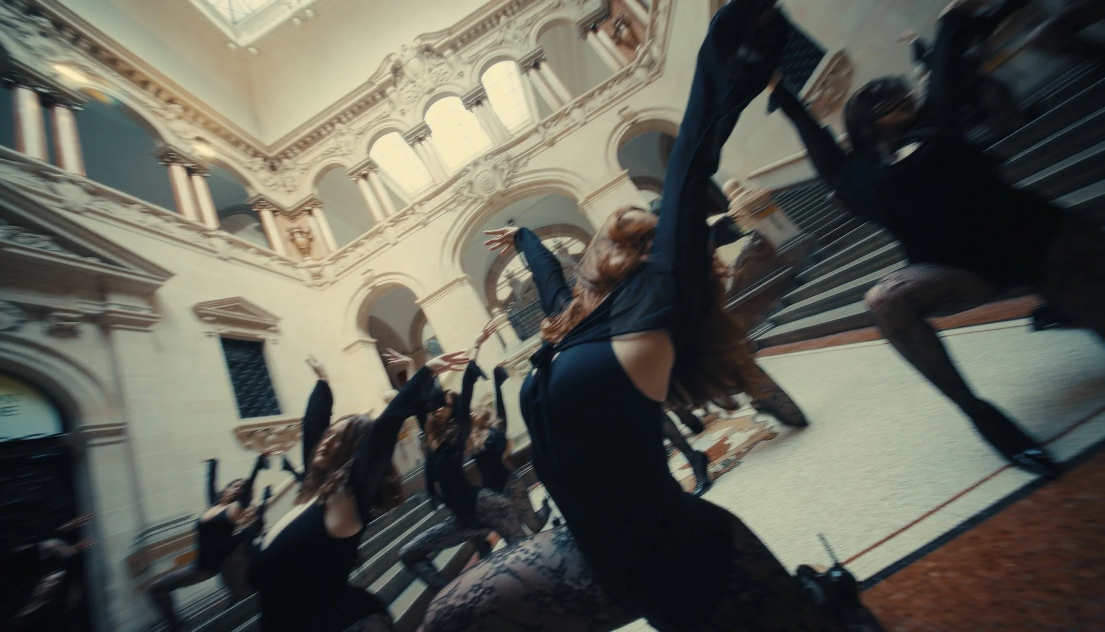 A group of people in black outfits performing a dance or fight scene on a grand staircase in an ornate, luxurious building with high ceilings and large windows.