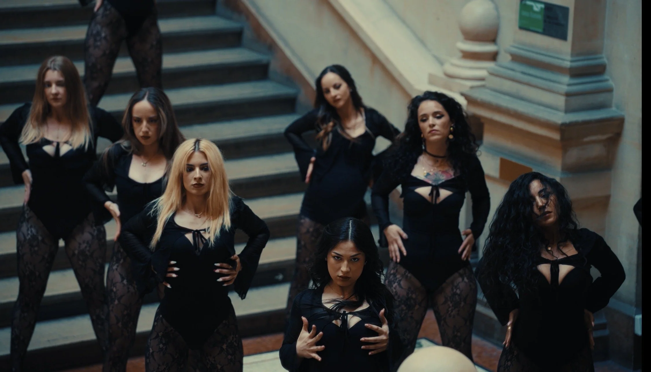 Group of women dressed in black outfits with lace and cutouts, posing on a staircase in an ornate interior setting.