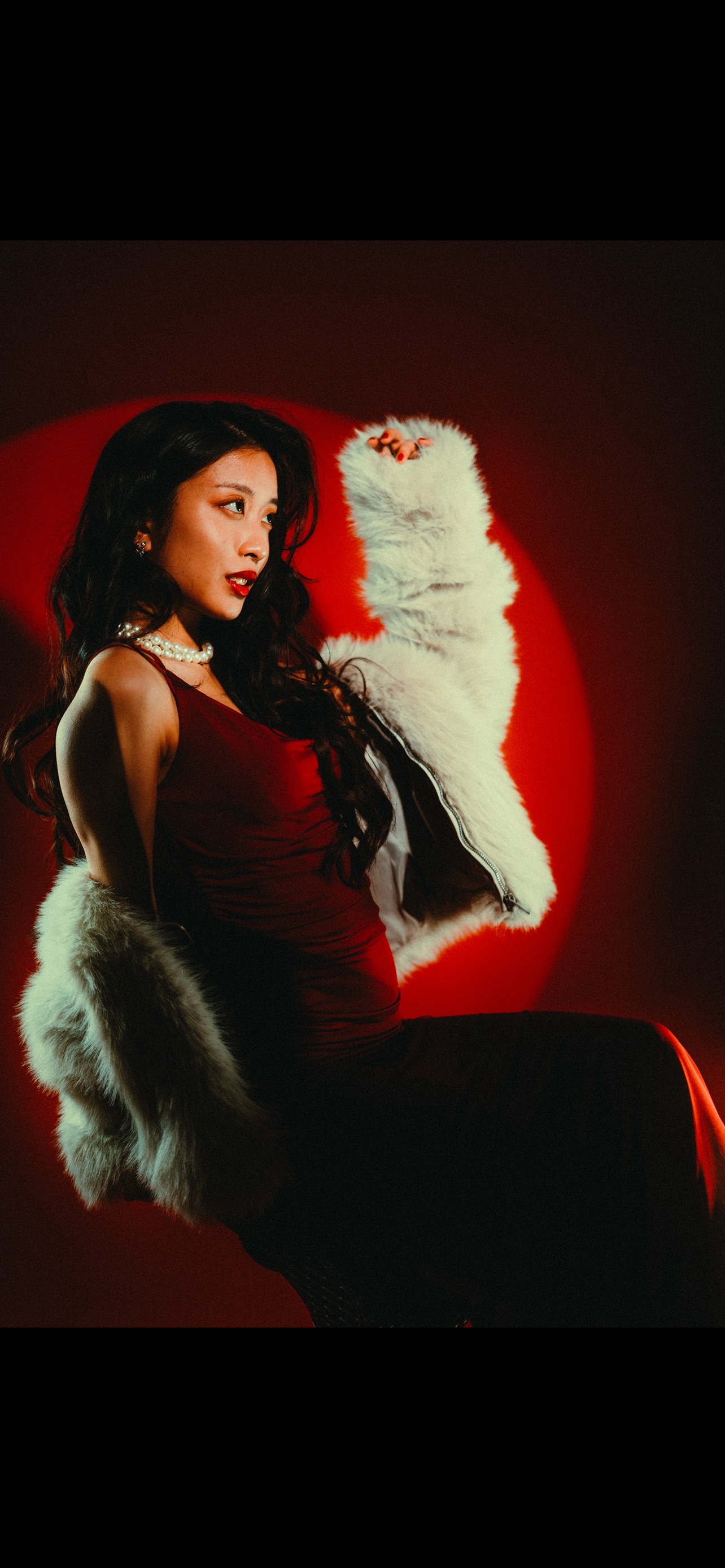 A woman with long dark hair wearing a red dress and pearl necklace, posing against a red circular backdrop, draped in a white fur coat with black trim.