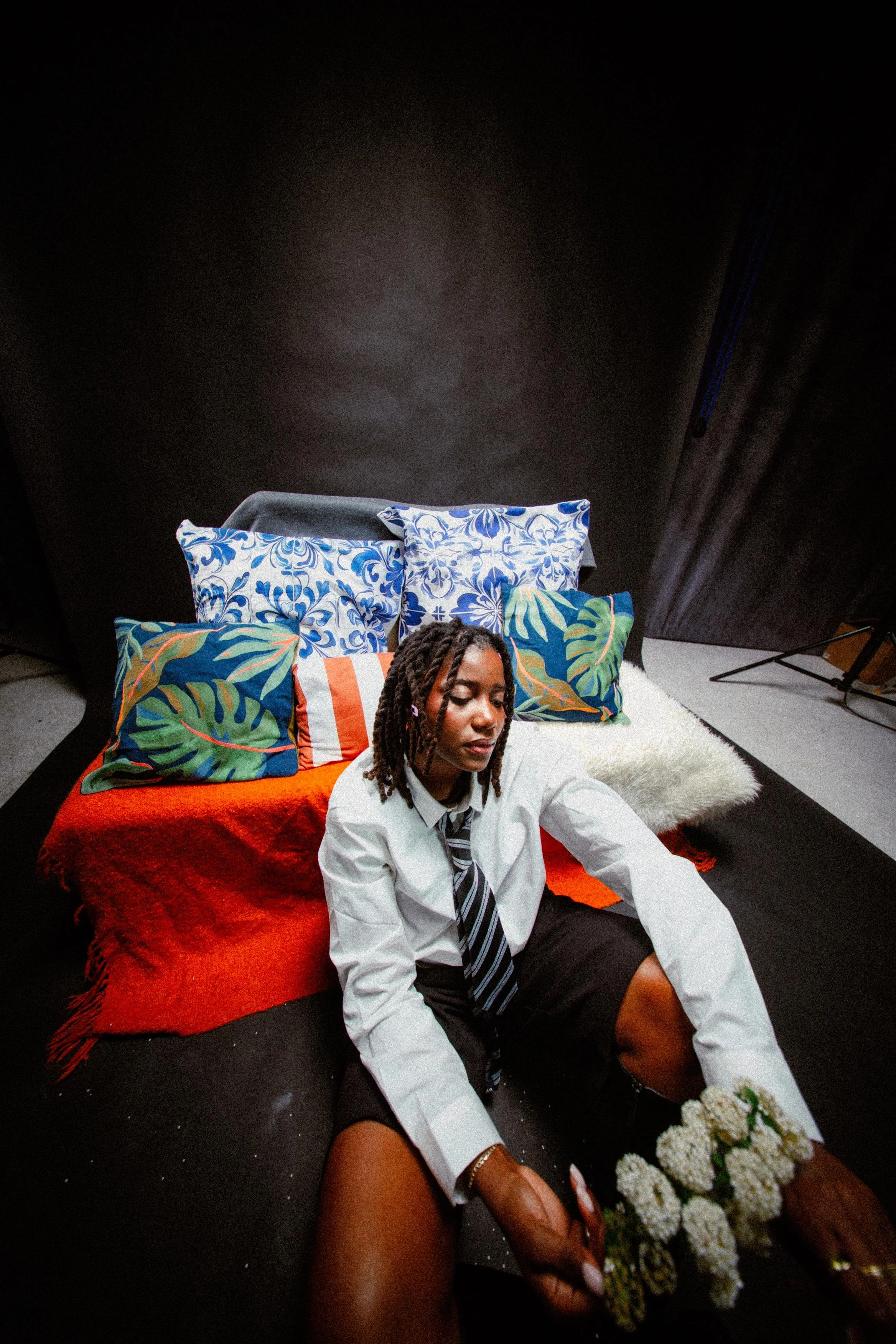 A woman with dreadlocks and a white shirt sitting on a black floor, holding a bouquet of white flowers, with multiple colorful pillows and a red blanket behind her.