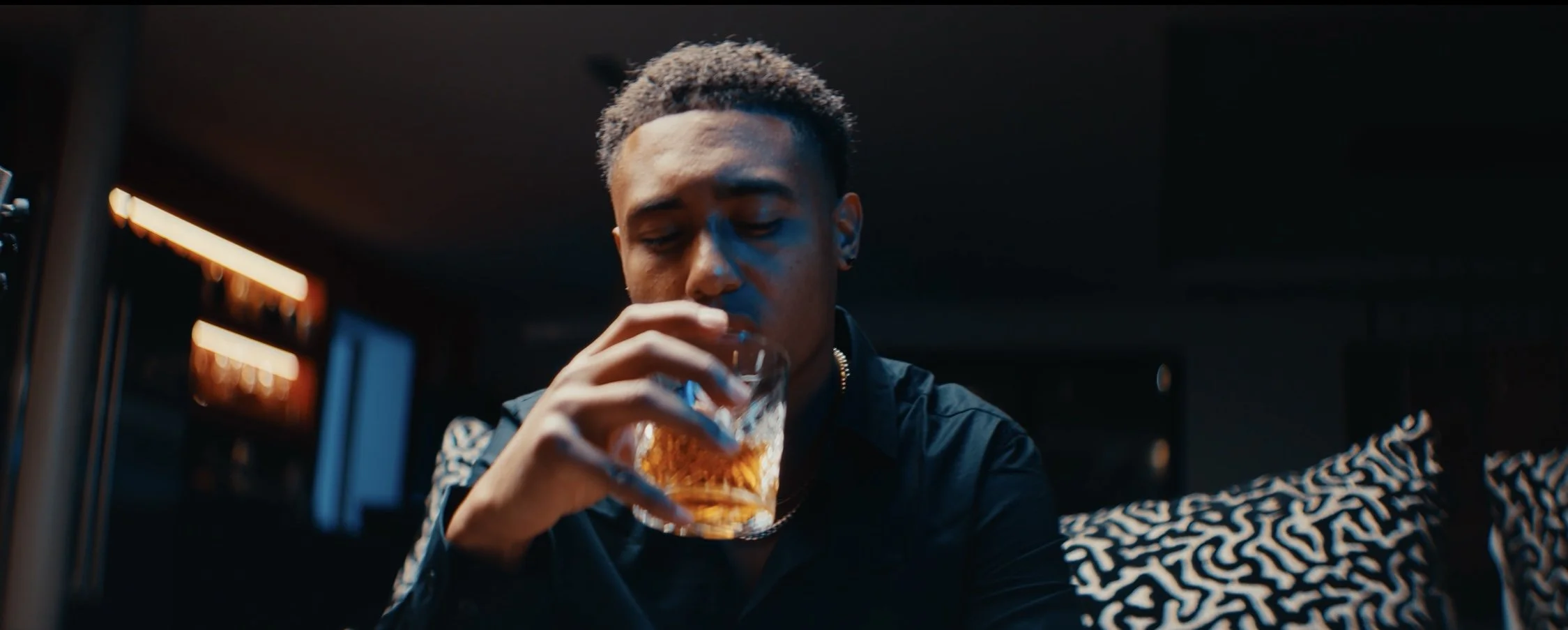 A young man with short hair, wearing a black jacket with a patterned sleeve, is sitting in a dimly lit room, drinking a glass of what appears to be whiskey or another amber-colored beverage.
