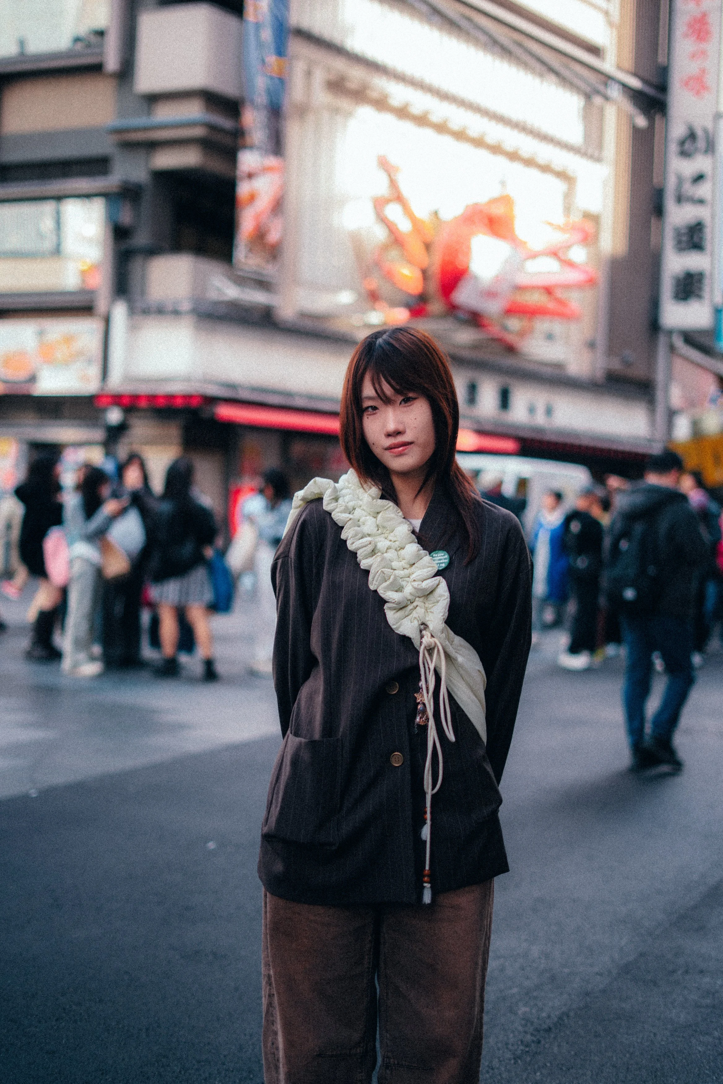 A young woman with dark hair and a white flower-shaped accessory around her neck, standing on a busy city street with blurred crowds, neon signs, and billboards in the background.