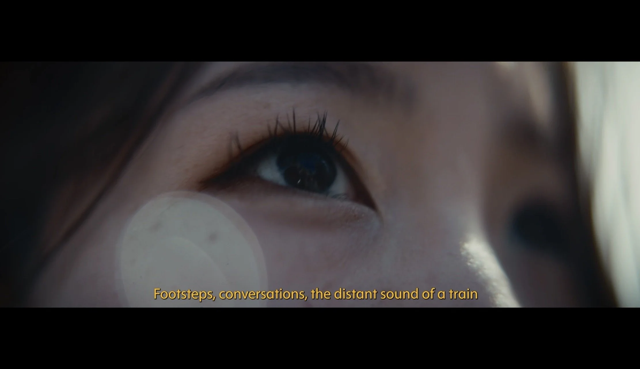 Close-up of a person's eye with yellow subtitles reading "Footsteps, conversations, the distant sound of a train."