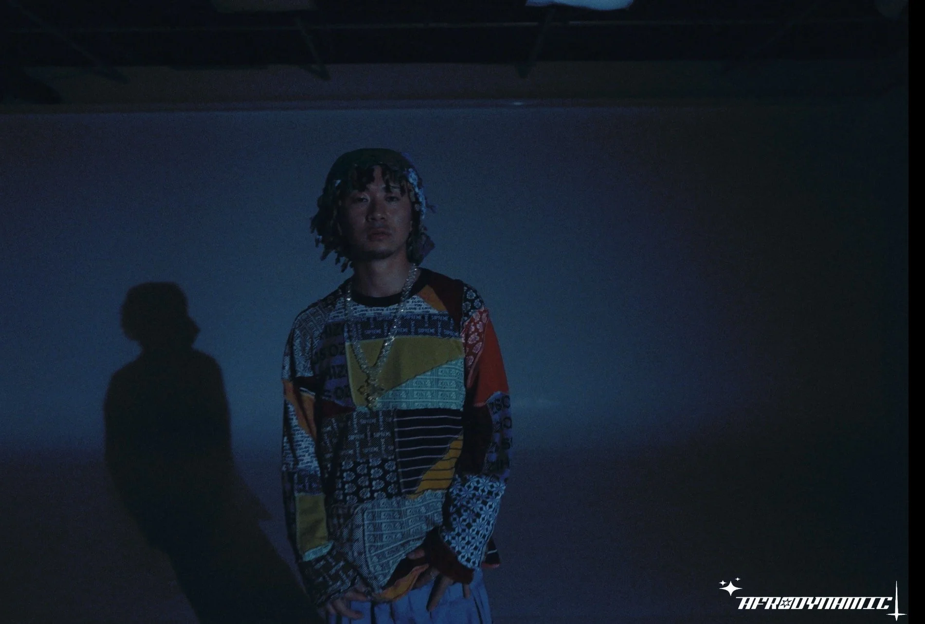 A person with dark, styled hair wearing a colorful patchwork shirt and a gold chain stands in a dark, misty environment with shadows casting behind him. The scene has a moody, atmospheric feel, and there's a logo in the bottom right corner reading 'A