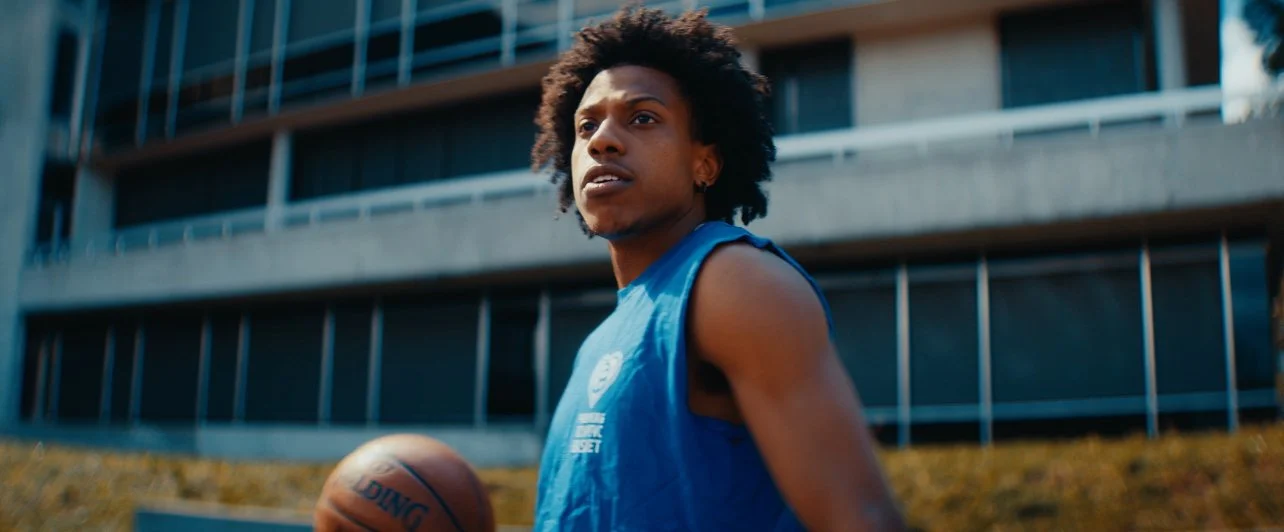 Young man with curly hair wearing a blue basketball jersey, holding a basketball, standing outside near a modern building.
