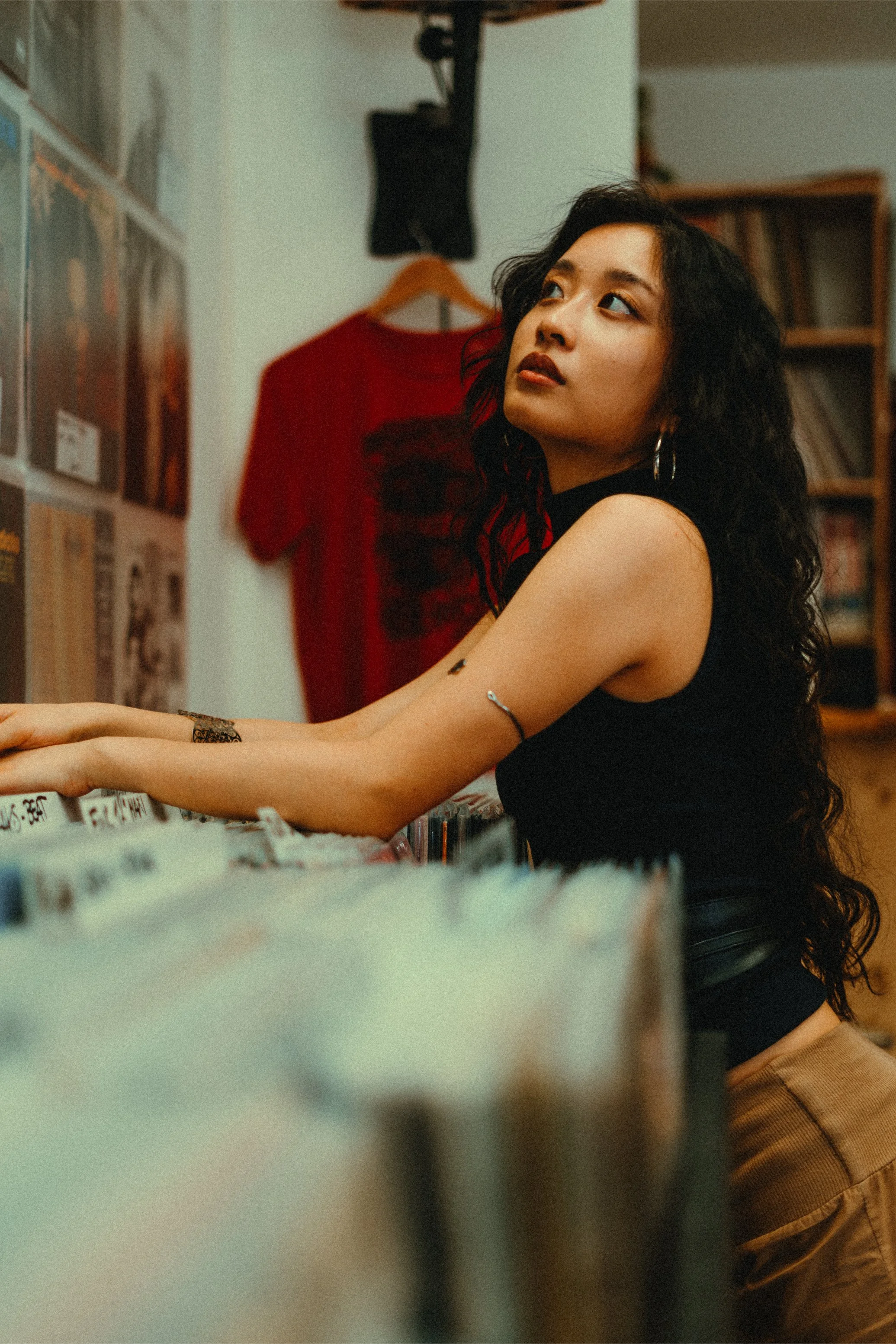 A woman with long, curly black hair and hoop earrings is browsing vinyl records in a record store, sitting at a table with records in front of her.