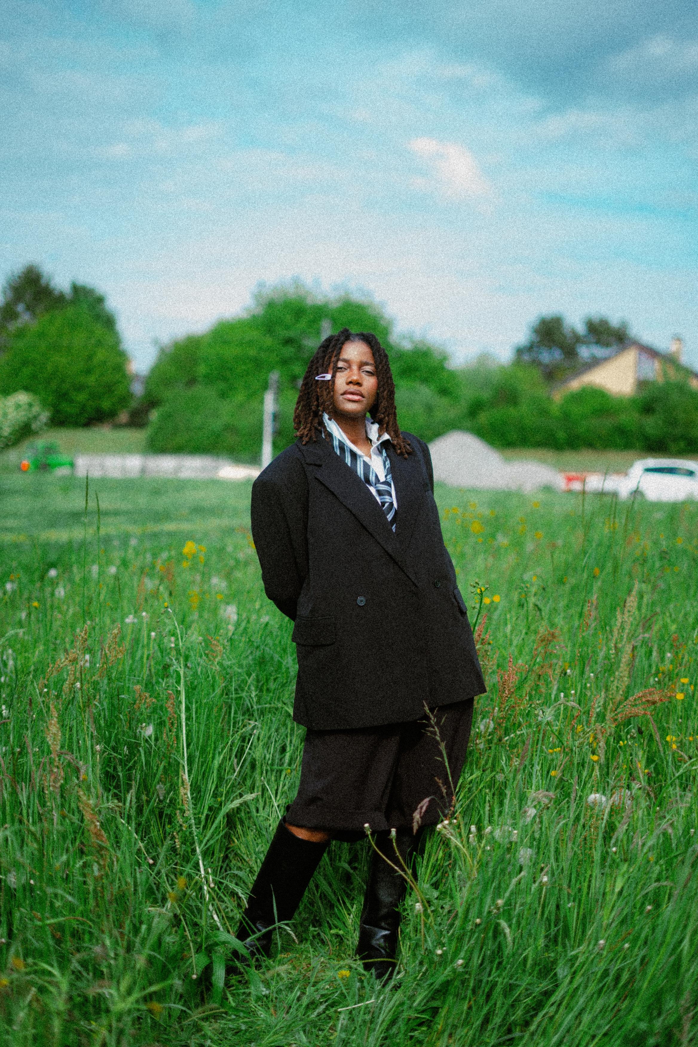 A woman wearing a black blazer and skirt, with a striped shirt, standing in a green field with tall grass and yellow flowers, with trees and houses in the background, under a partly cloudy sky.