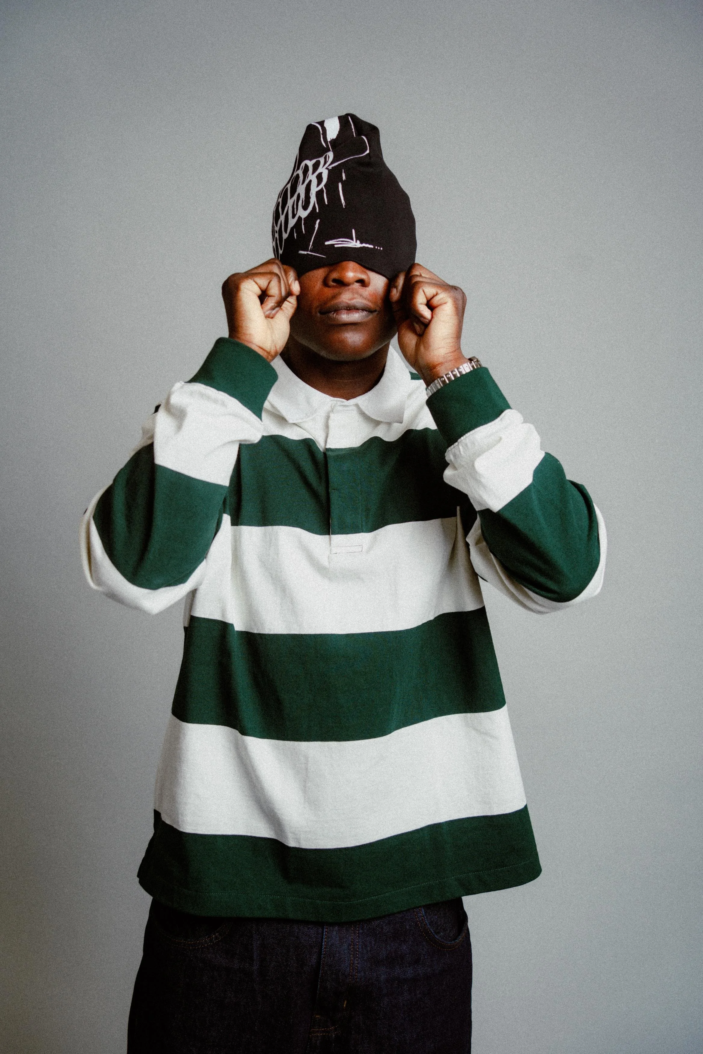 A person wearing a green and white striped pullover with a watch on their left wrist and dark jeans, with a black beanie covering their eyes, pulling the beanie down with both hands, against a plain grey background.