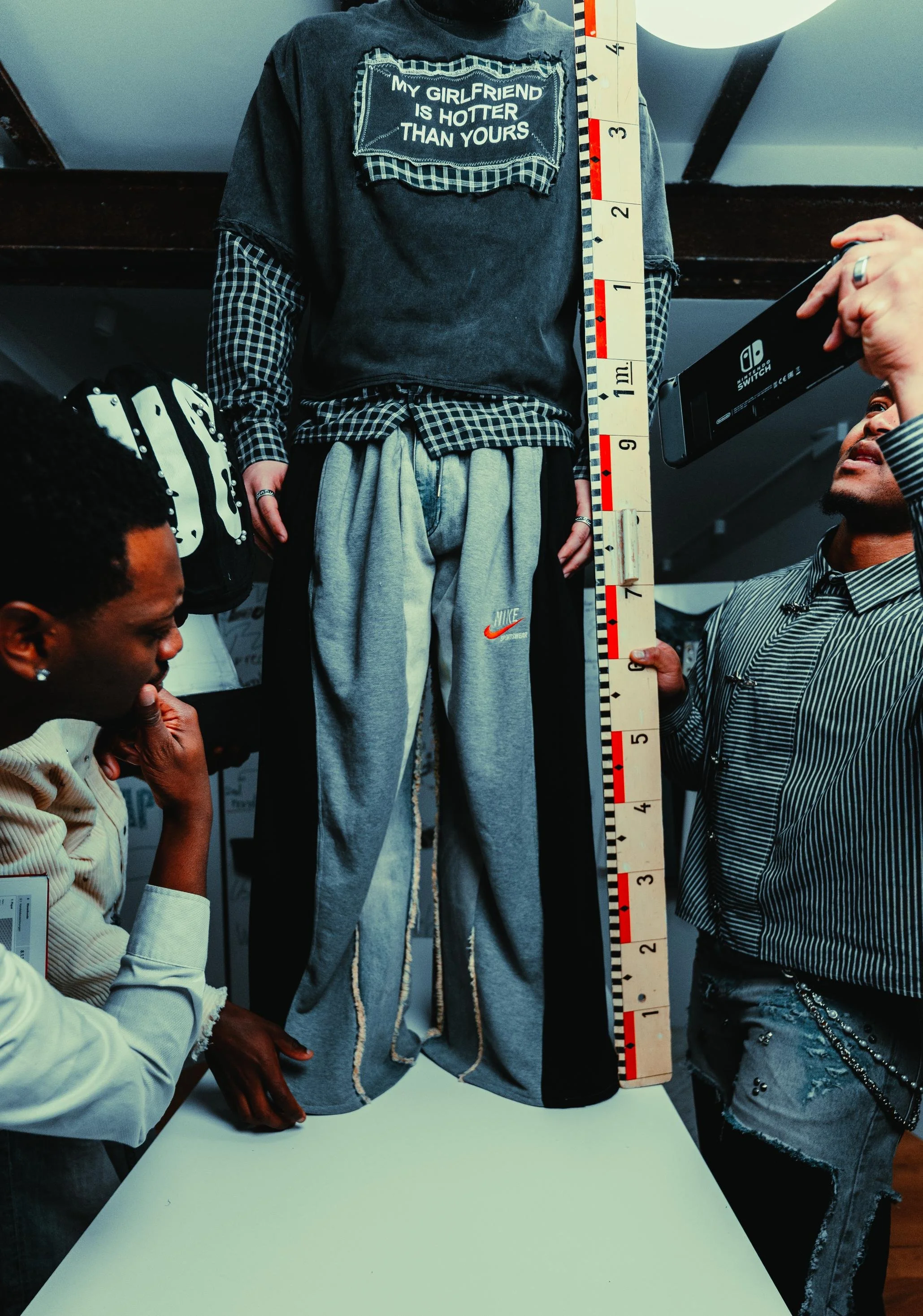 A person wearing a black T-shirt with a patch that says 'My girlfriend is hotter than yours,' standing next to a measuring stick with black and red markings, while two others are observing. One is taking a photo with a smartphone, and another is in a beige jacket with braided embellishments.