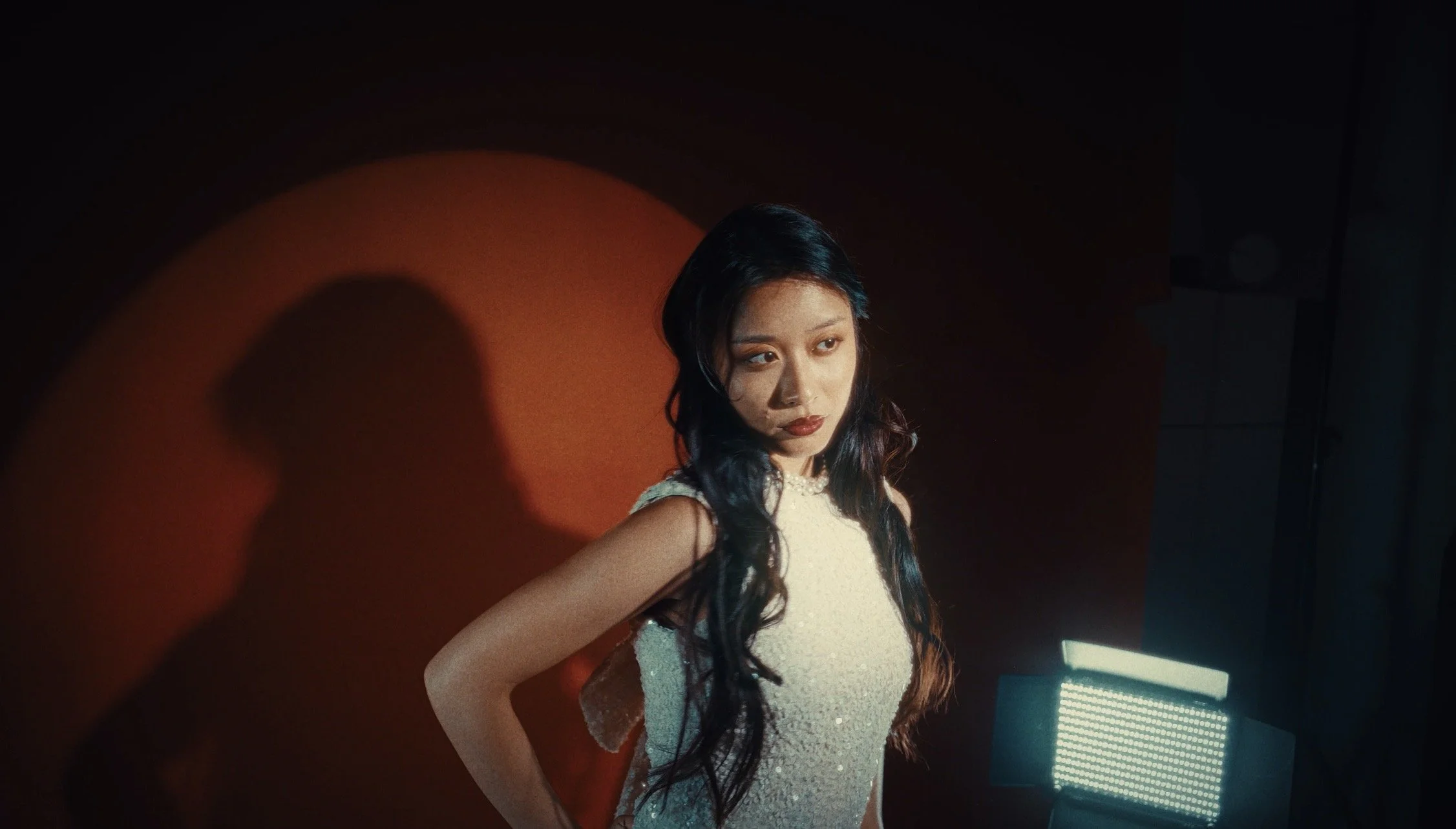 A young woman with long black hair wearing a white dress standing in a dimly lit room with a dark background and a red wall.
