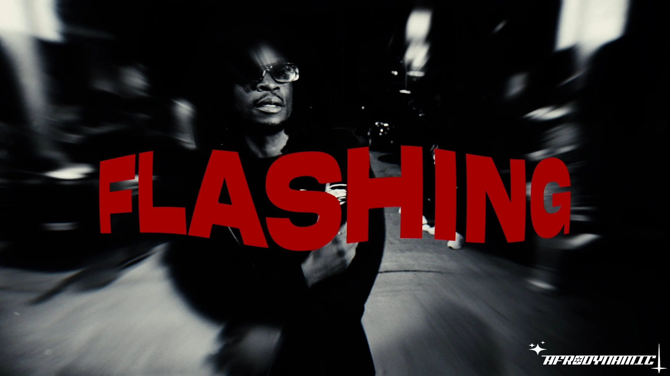 A person wearing glasses and black clothing, with a blurred background, overlaid with the red text 'FLASHING'. The image has a dark, high-contrast, stylized appearance with a dynamic, streaked effect.