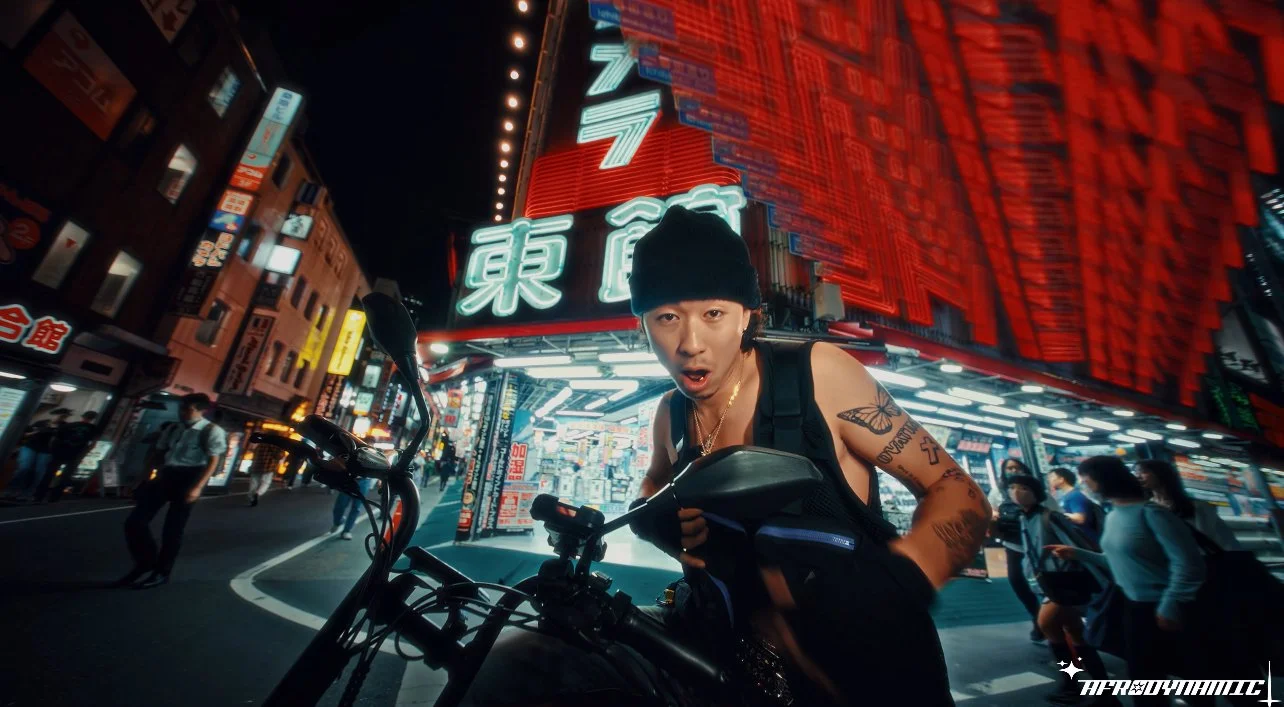 A person with tattoos and wearing a black beanie and sleeveless top standing next to a bicycle on a busy city street at night, illuminated by bright neon signs in Asian characters.
