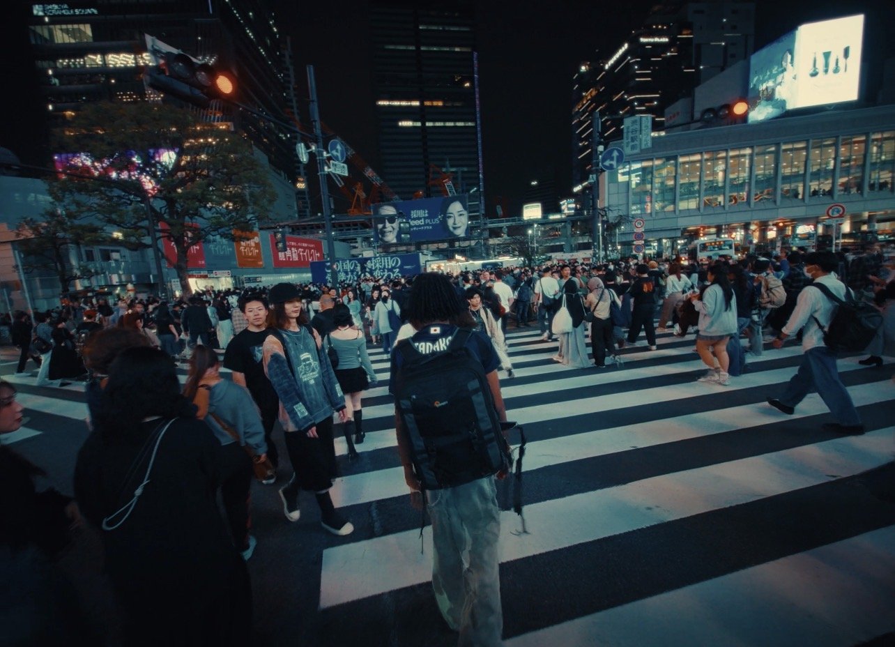 A crowded crosswalk at night in a busy city, with numerous pedestrians walking across, illuminated billboards, and modern buildings
