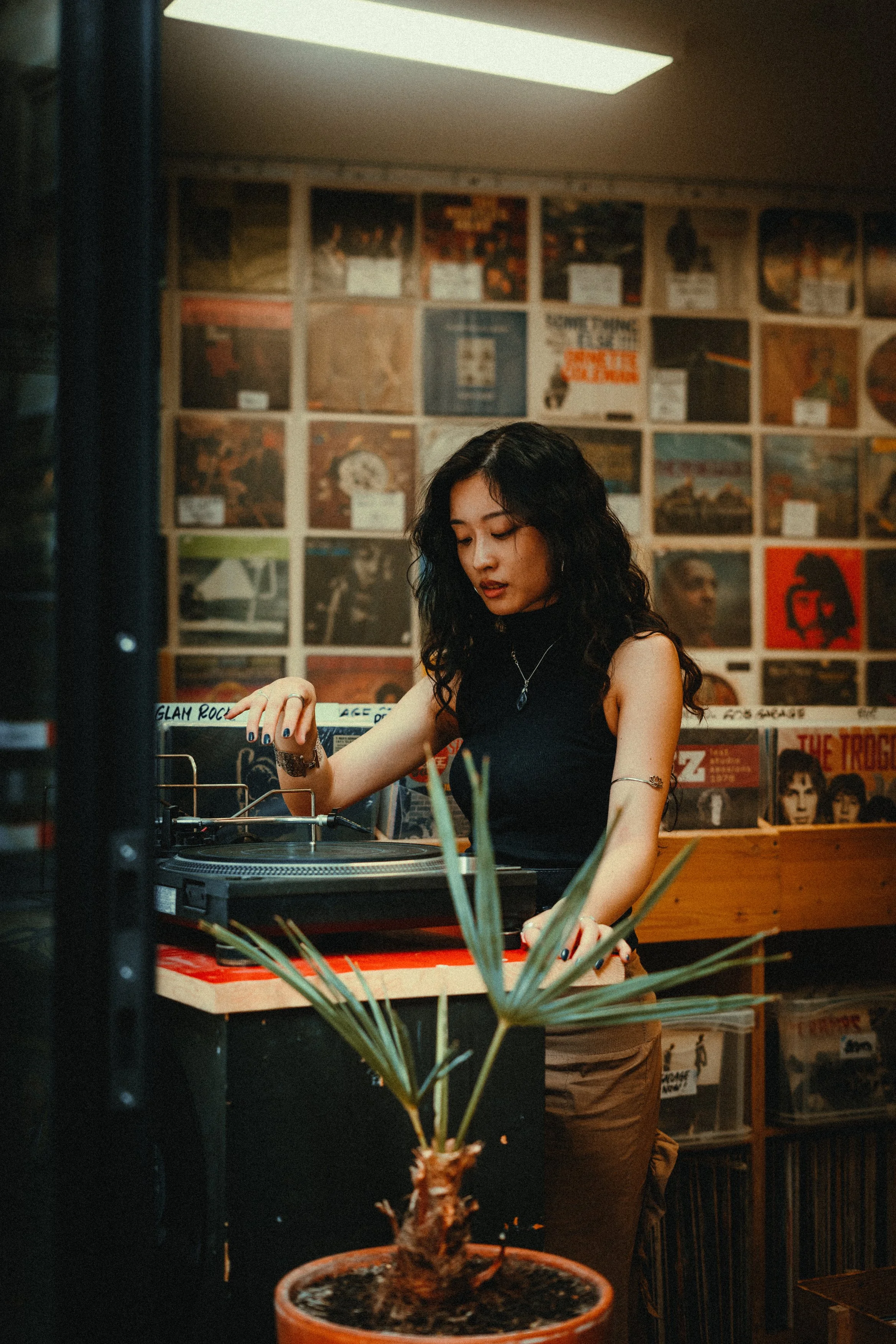 A woman with long dark curly hair wearing a black sleeveless top and silver jewelry, appears to be DJing or playing music on a turntable inside a record store or cafe, with vinyl records and posters on the wall behind her, and a potted plant in the f